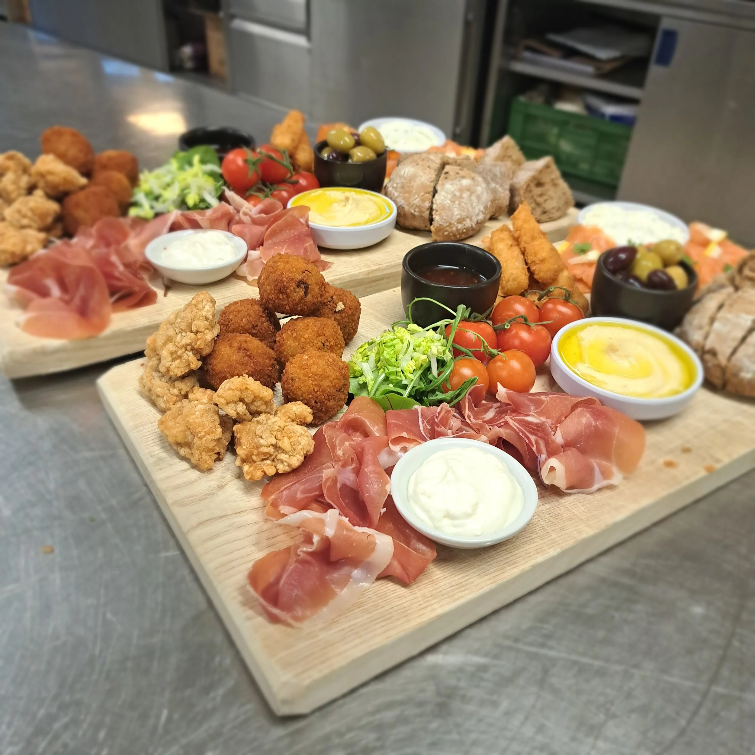 Assortiment van vleeswaren, zoals ham en salami, samen met rijpe tomaten, groene olijven, gefrituurde kipnuggets, brood, kruidenboter, dipsauzen en verse sla op houten planken.