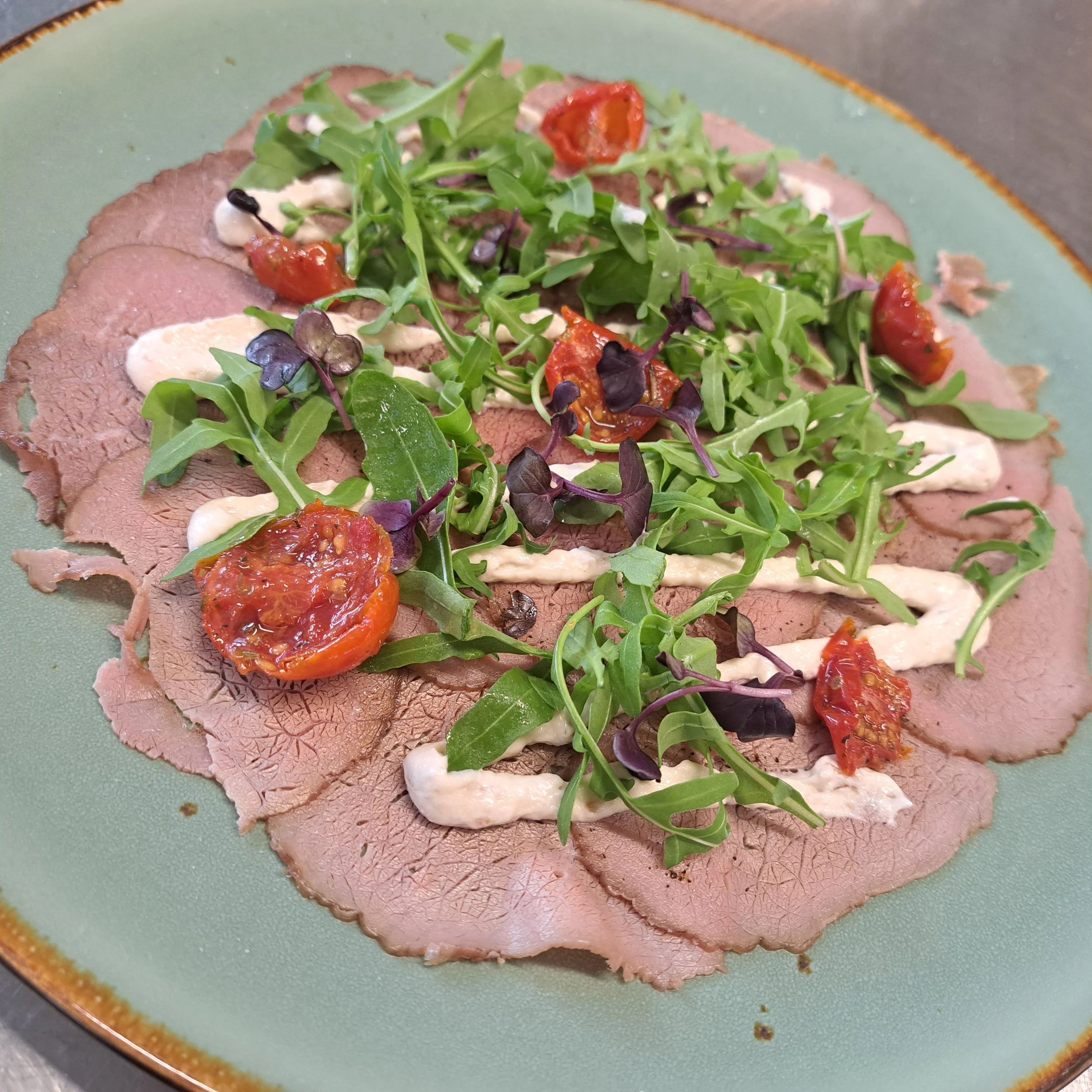 Een bord met plakjes gekookt kalfsvlees, bedekt met gemengde salade en cherrytomaatjes.