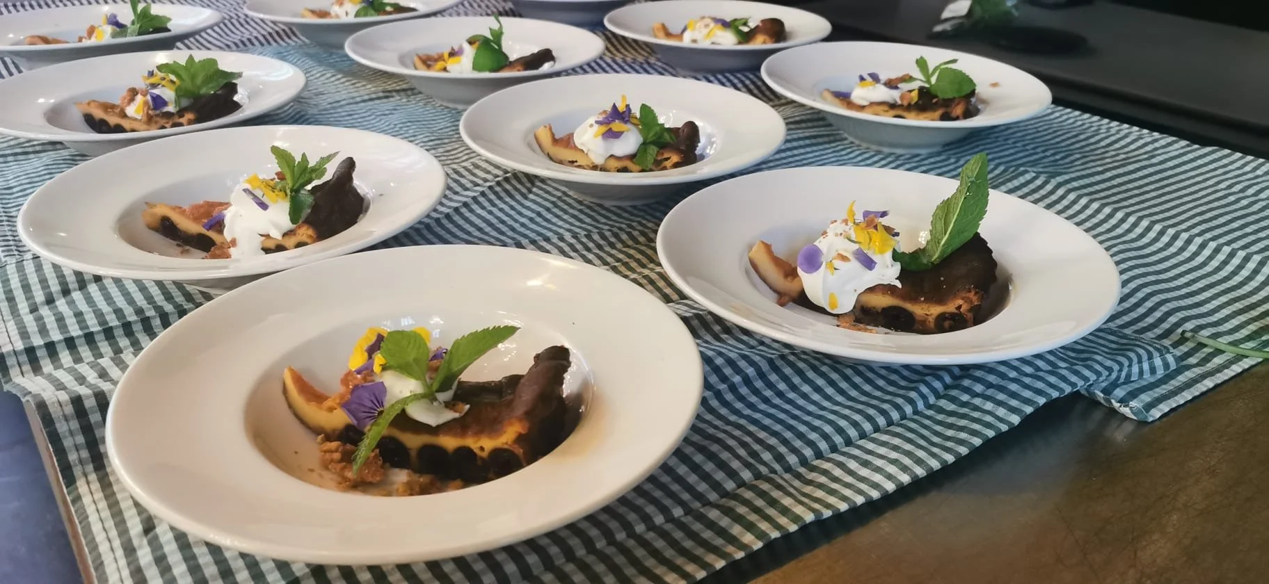 Tien witte borden met een stuk chocoladetaart, groen muntblad, slagroom, en eetbare bloemen, uitgestald op een tafel met een blauwe en witte geruite tafelkleed.