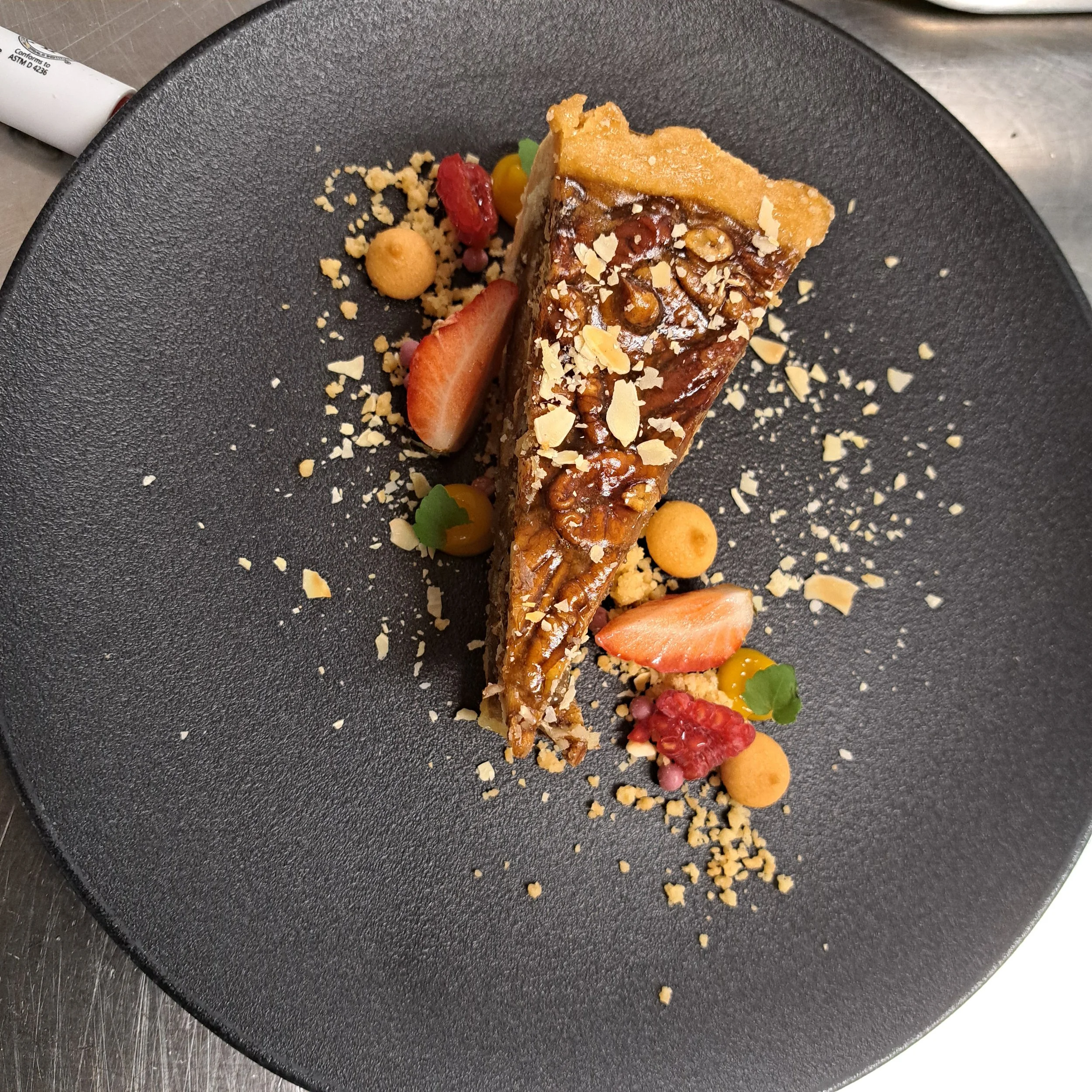 Een stuk taart met karamelsaus en noten op een zwarte bord, versierd met aardbeien en crumbles.