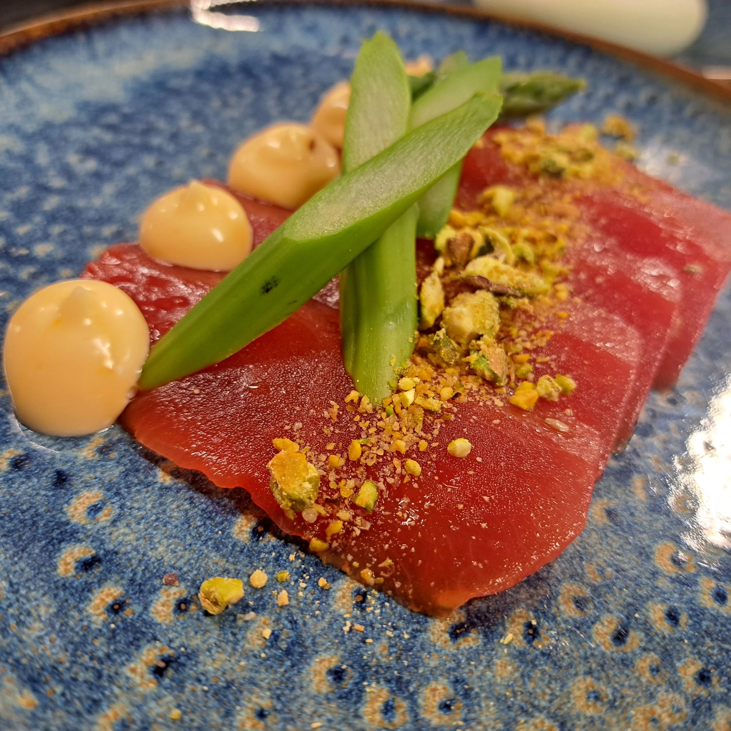 Close-up van een gerecht met gerookte zalm, pistachenoten, groene asperges, en een saus op een blauwe bord.