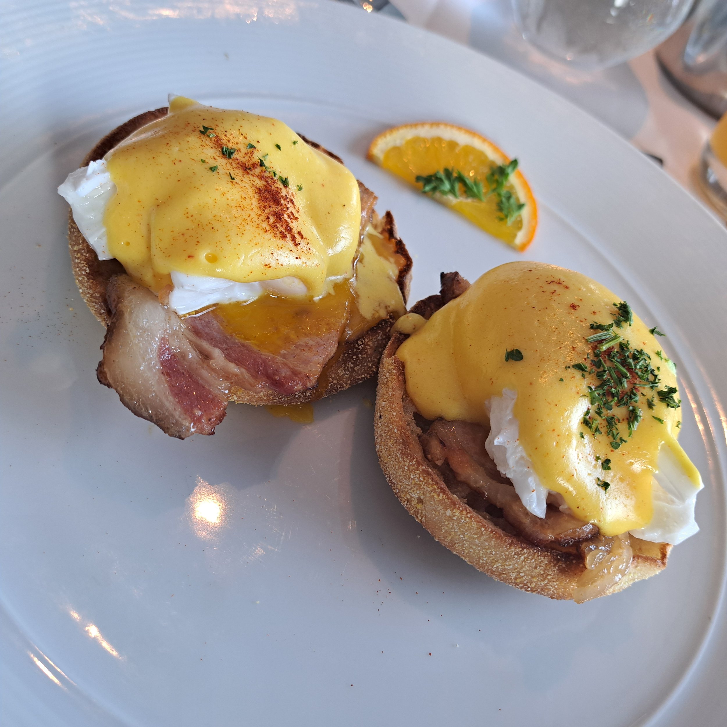 Een bord met Eggs Benedict, bestaande uit een gebakken eidooier, ham, en Hollandaisesaus, geserveerd met een stukje brood en een schijfje sinaasappel met peterselie.