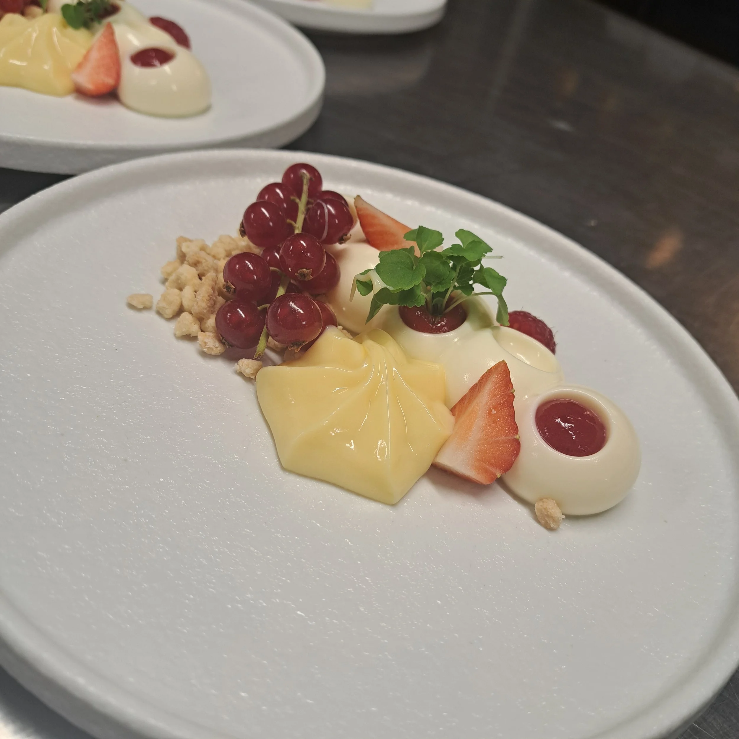 Een bord met een dessert dat bestaat uit rood fruit, een stuk aardbei, groene kruiden, crumbles en saus, geserveerd op een wit bord.