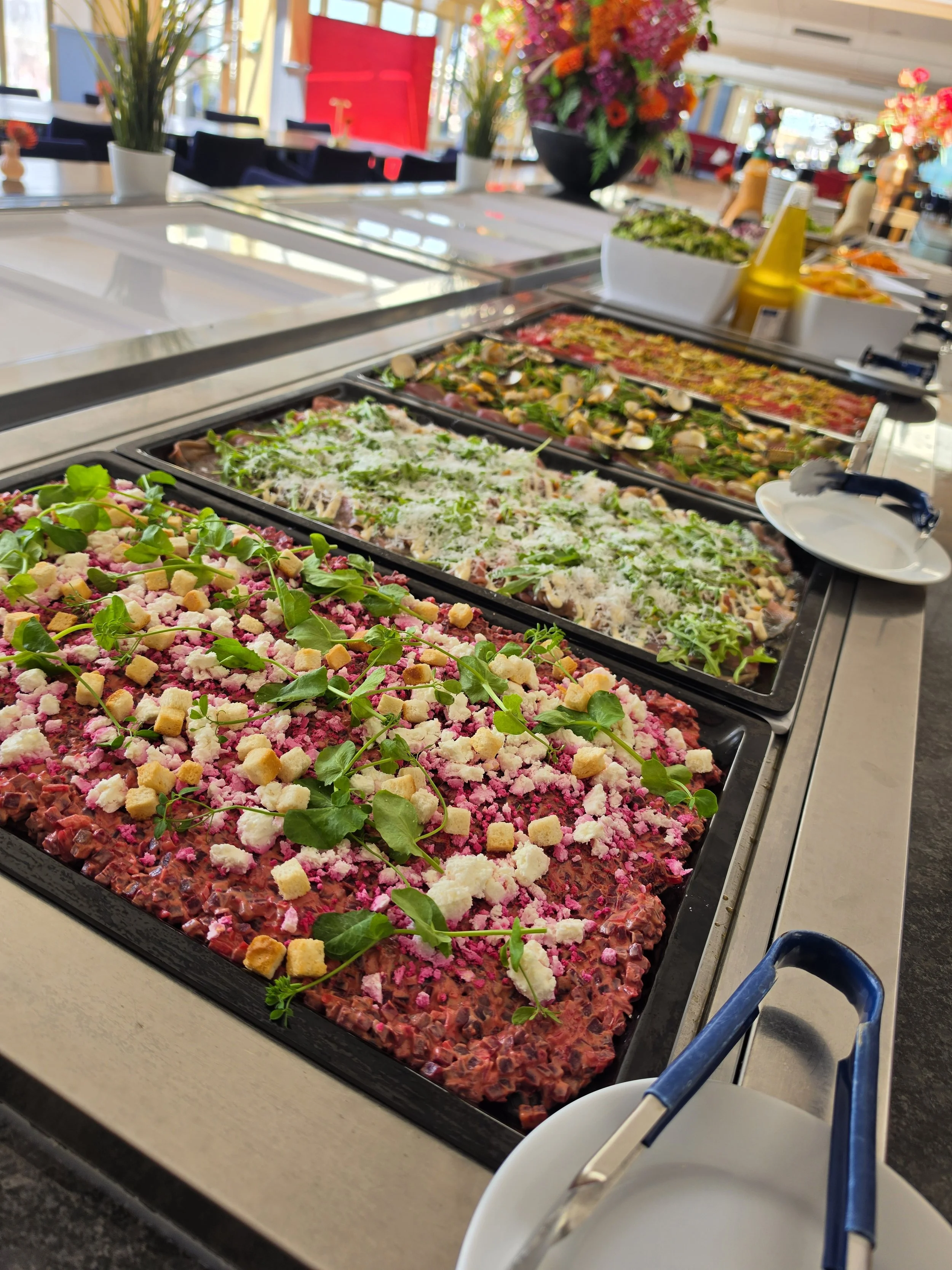 Saladebuffet met verschillende soorten salade in grote bakken op een tafel, decoraties en bloemen op de achtergrond.