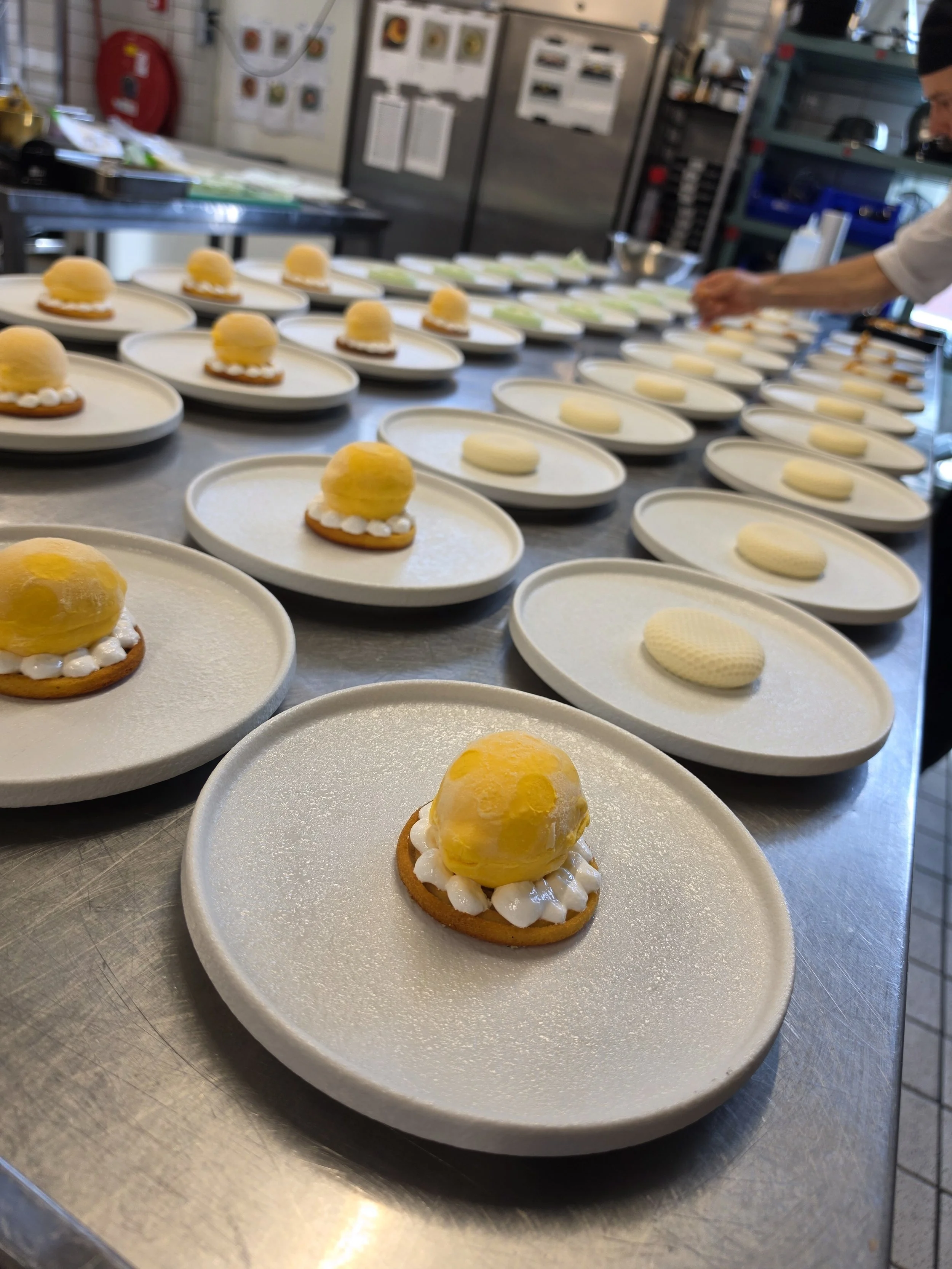 Een rij platters met desserts, waarbij de voorste een dessert met een gele bol bovenop, crème en een koekje. De achtergrond toont meerdere identieke desserts en een persoon die er aan werkt in een keukensetting.