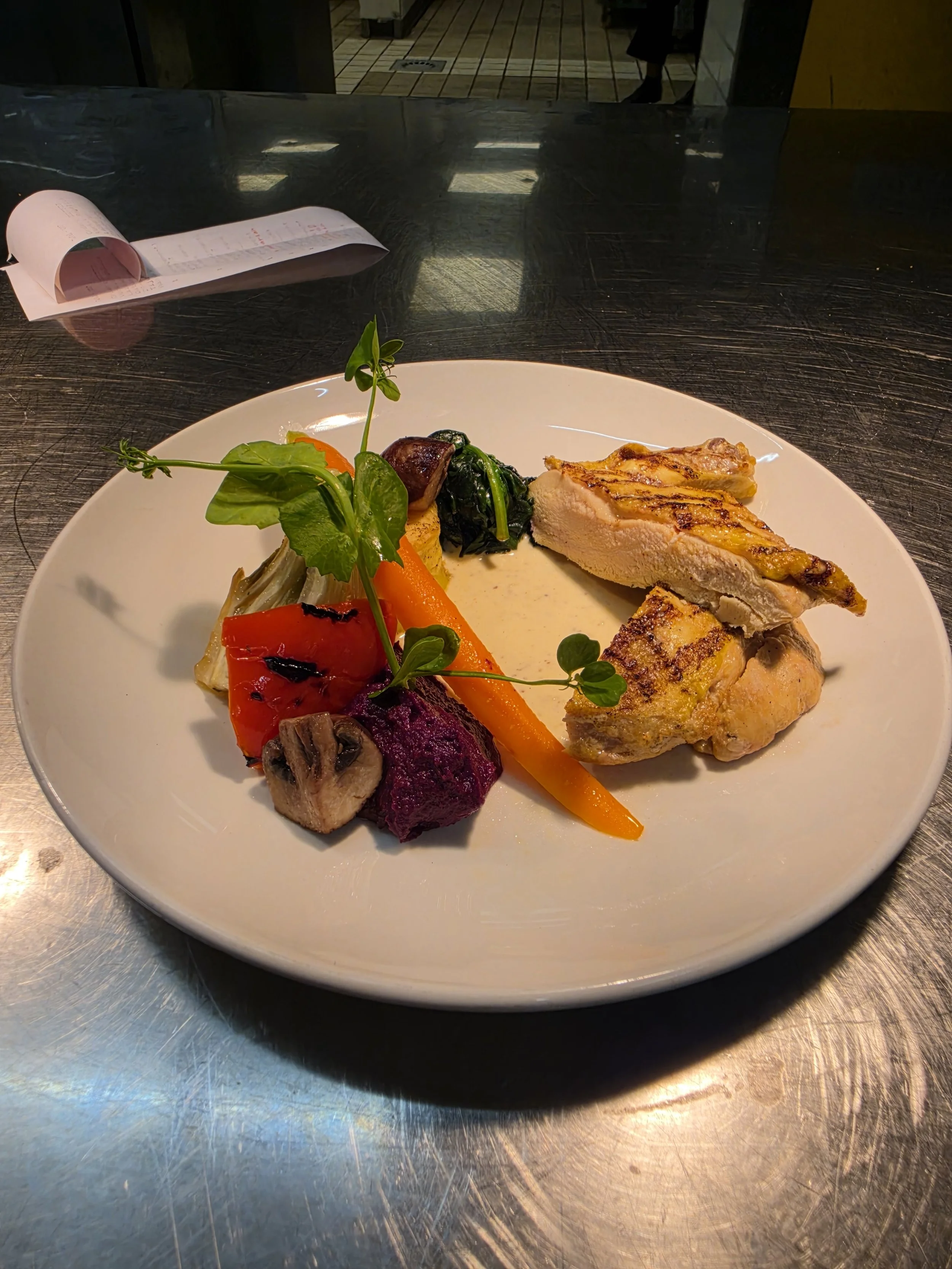 Gerecht met groenten en gevogelte op een wit bord, geplaatst op een metalen tafel, met een kassabon op de achtergrond.