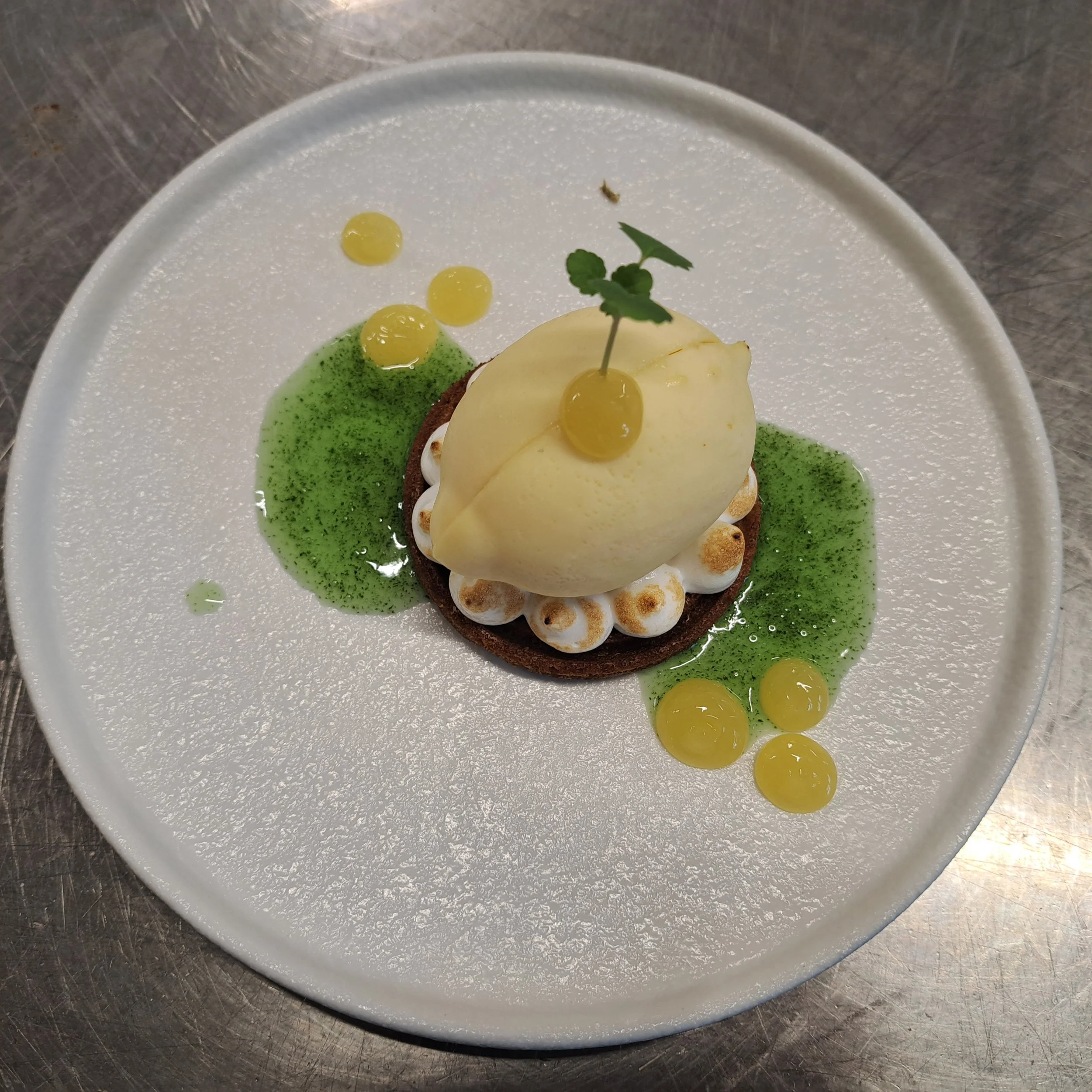 Een wit bord met een dessert dat bestaat uit een taartbodem met slagroom en een stuk peer er bovenop, versierd met een takje munt en kleine gele gelletjes, op een metalen tafel.