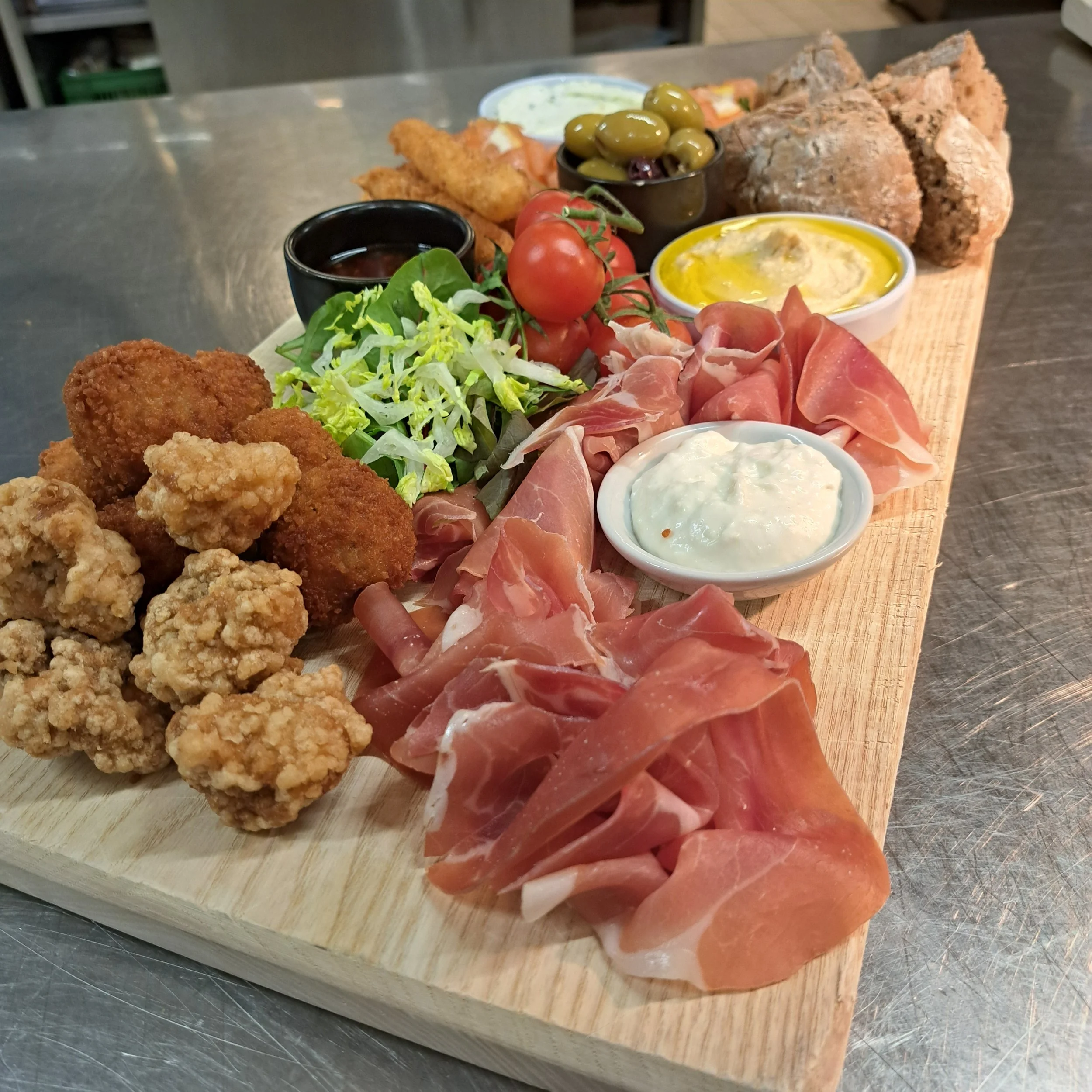 Levensbord met diverse hapjes zoals gebraden kip, vleeswaren, groene olijven, cherrytomaten, dipsauzen en salade, op houten plank.