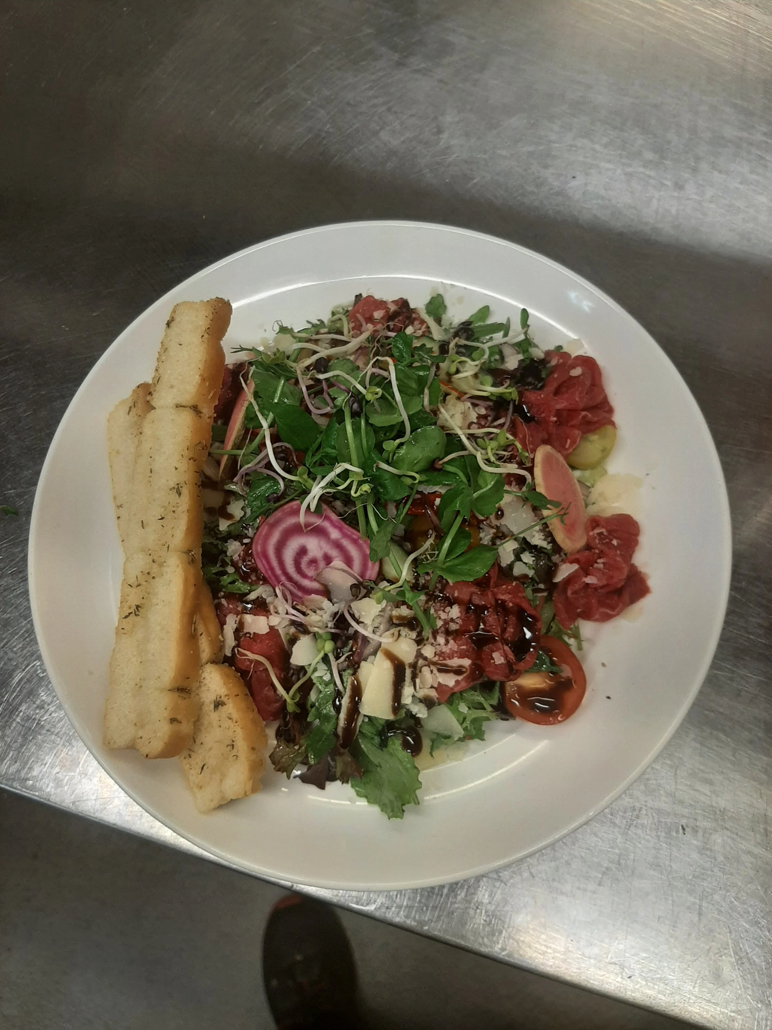 Een salade met tomaten, radijsjes, komkommer, gemengde sla en een kaasrol, geserveerd op een wit bord.