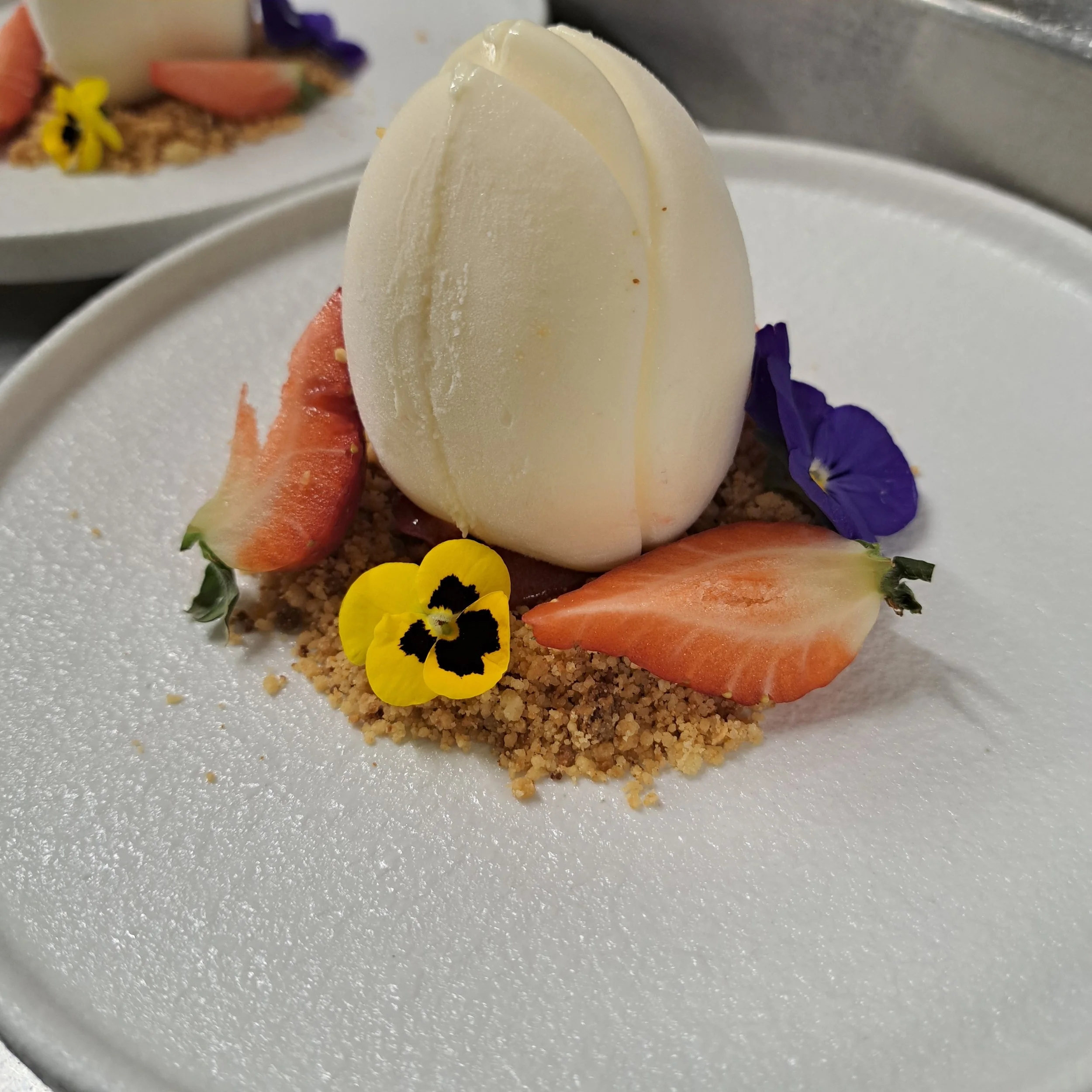 Een wit chocolade-ei op een bord met vers fruit, bloemen en crumble eromheen.