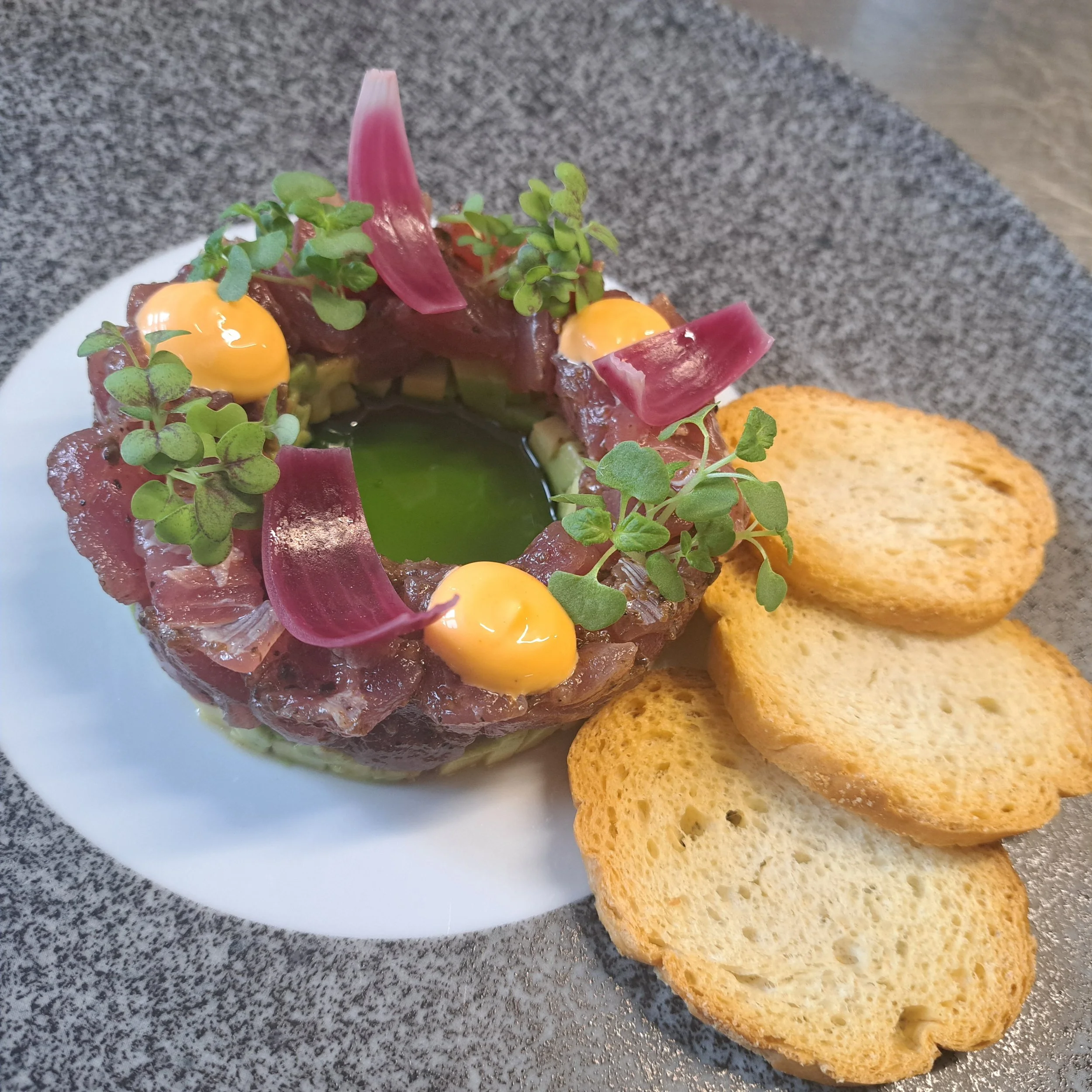 Een bord met tartaar, roze sieruien, groene microgroenten en een saus. Naast wat geroosterde toast.