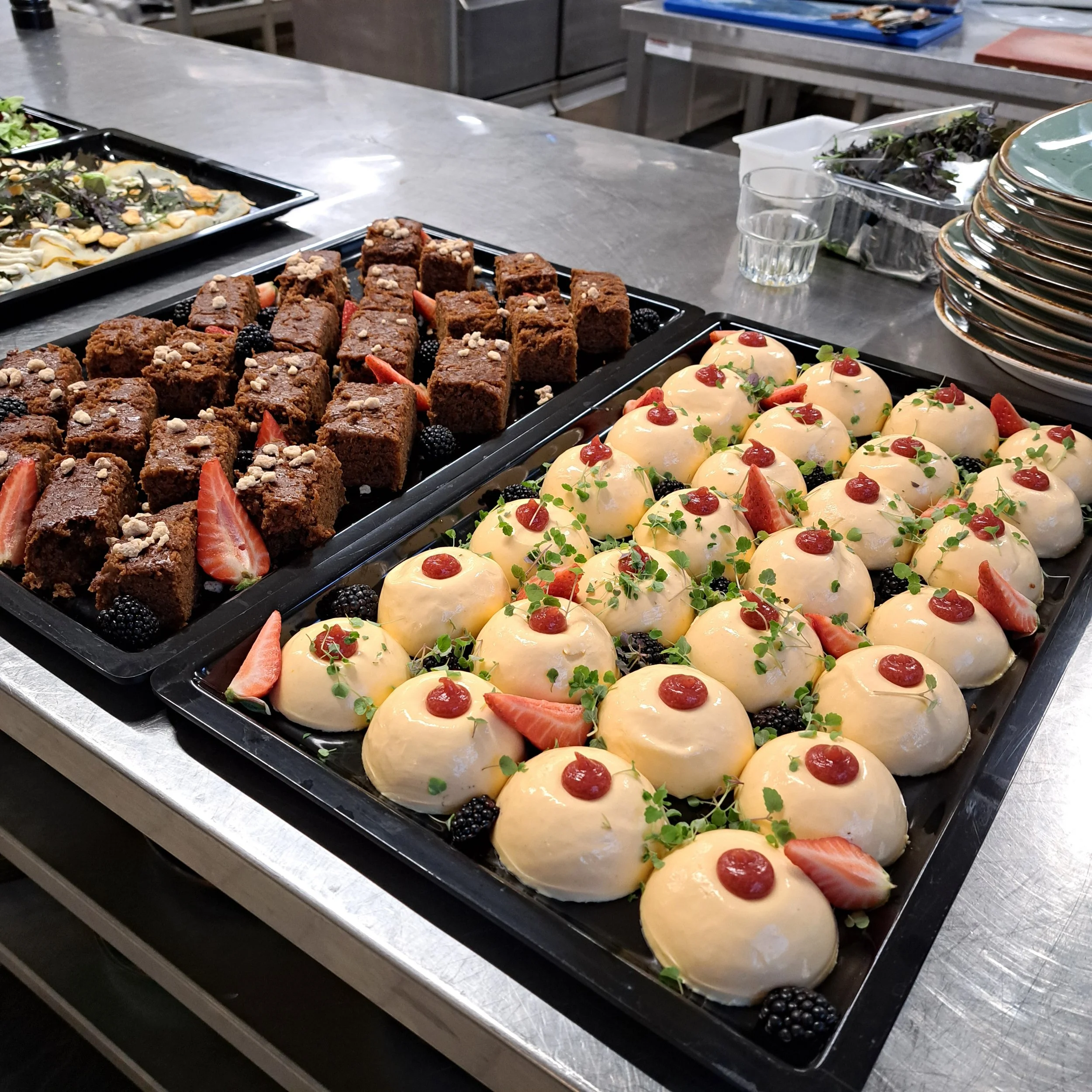 Een gerecht met kleine cake-achtige desserts versierd met aardbeien, bramen, groen en rode kersen, op een zwart dienblad. Daarnaast liggen er andere taarten en salades op een keukenblok.