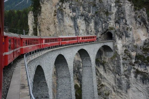 Roter Zug fährt über ein steinernes Viadukt in einer felsigen Landschaft in Graubünden.