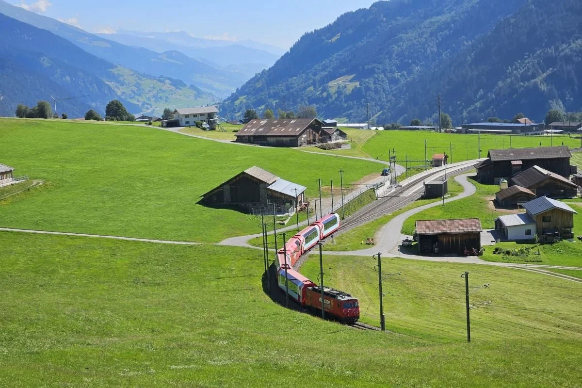 Roter Zug fährt in einer Kurve durch eine grüne Hügellandschaft mit Bauernhäusern, Haltepunkt und Fahrleitungsmasten; Berge im Hintergrund und blauer Himmel.