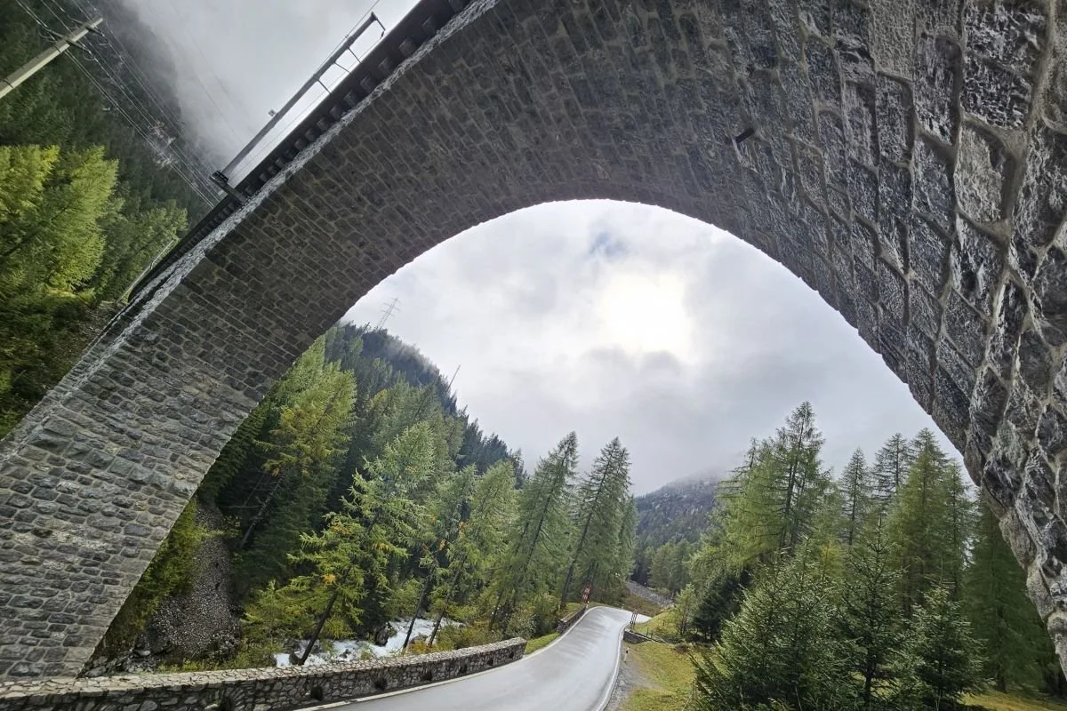 Steinbogenbrücke über einer kurvigen Strasse in einem bewaldeten Alpental, mit Nebel und diffuser Sonne.
