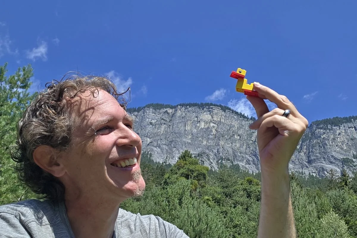 Hand hält eine kleine gelbe LEGO‑Ente im Aussenbereich, mit unscharfem Naturhintergrund.