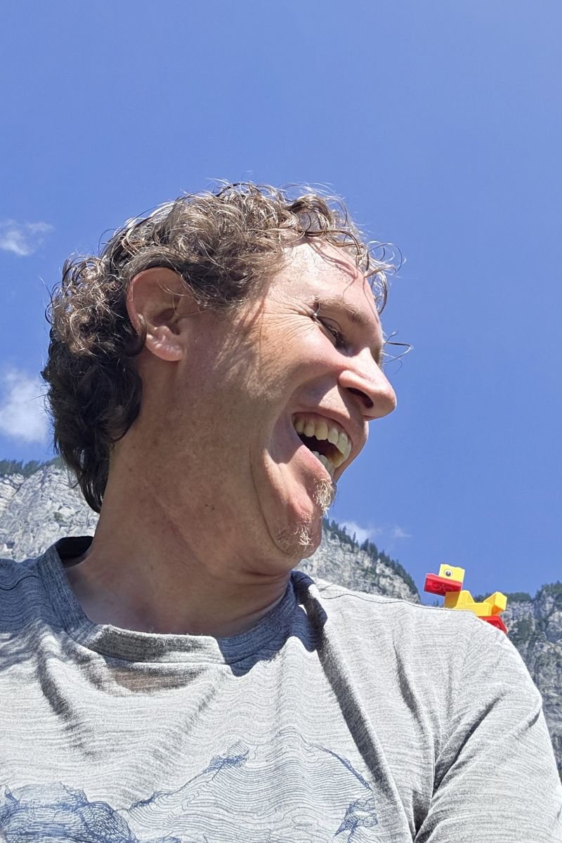 Person im Freien vor Fels- und Himmelhintergrund; am Bildrand eine kleine gelbe LEGO‑Ente sitzend auf der Schulter der Person.
