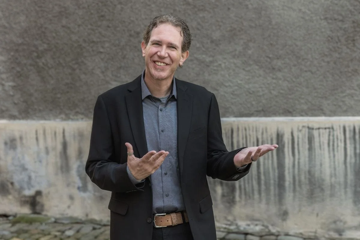 Person im Oberkörperausschnitt vor einer neutralen Wand, mit geöffneten Händen in gestischer Haltung.