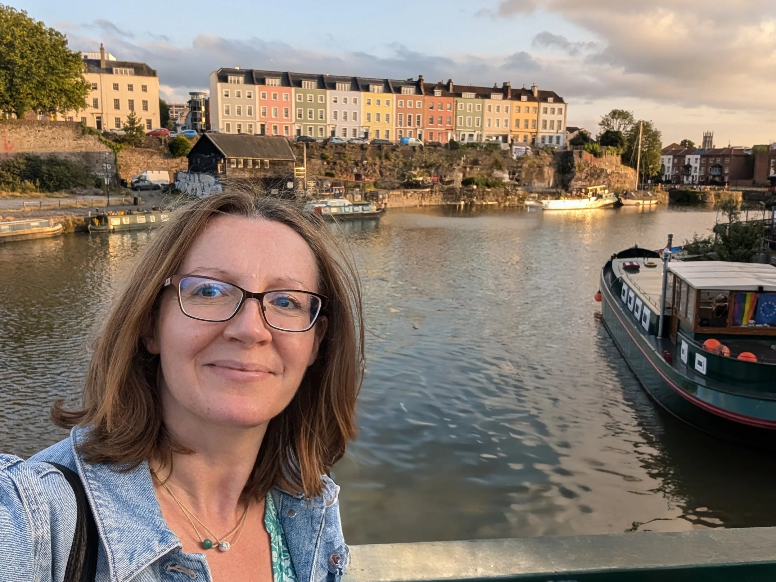 A woman with glasses taking a selfie by a river at sunset, with colorful houses and boats in the background.