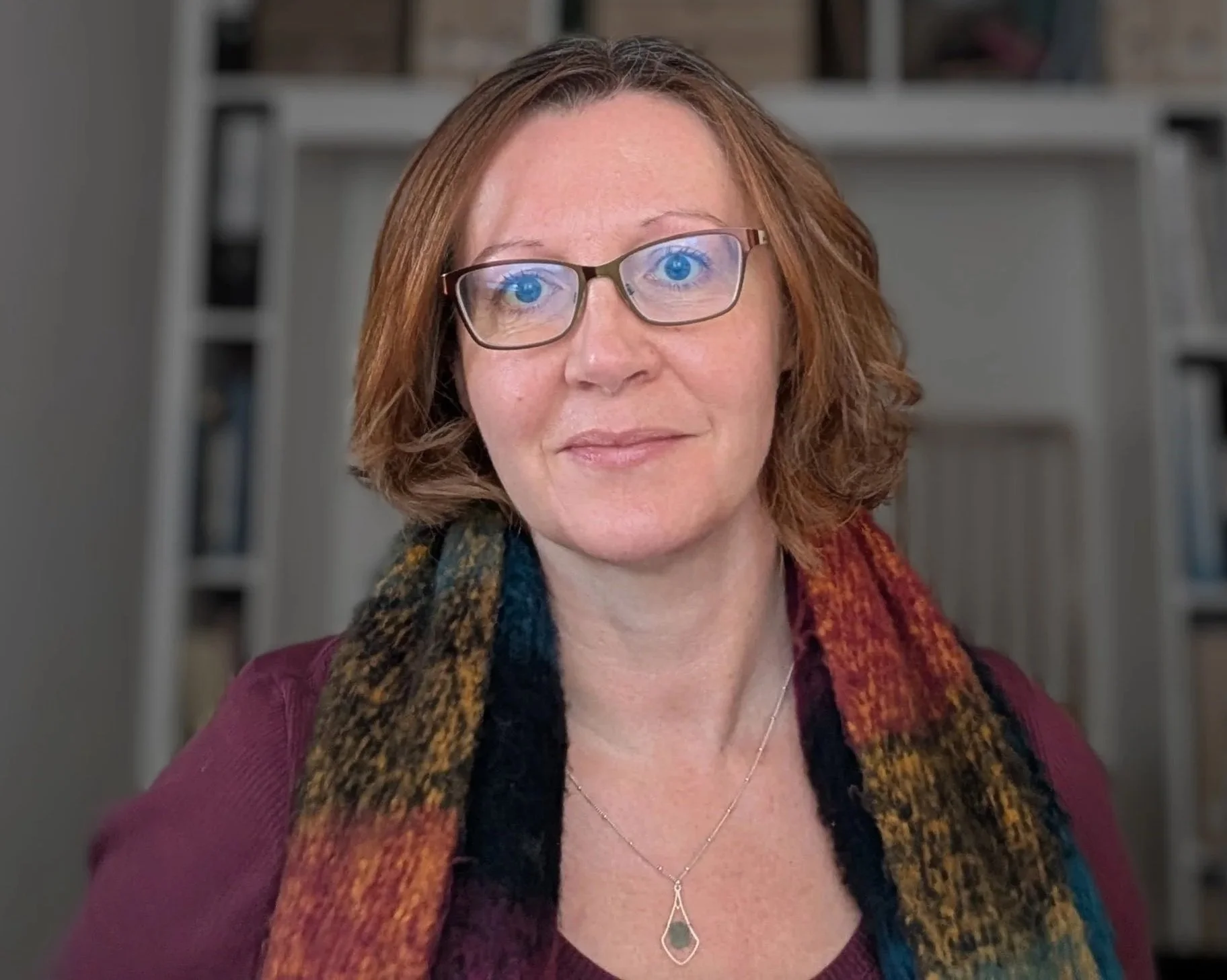 A woman with shoulder-length reddish-brown hair, wearing glasses, a burgundy top, a multicolored scarf, and a teardrop-shaped necklace, smiling slightly in front of a collection of white bookshelves.
