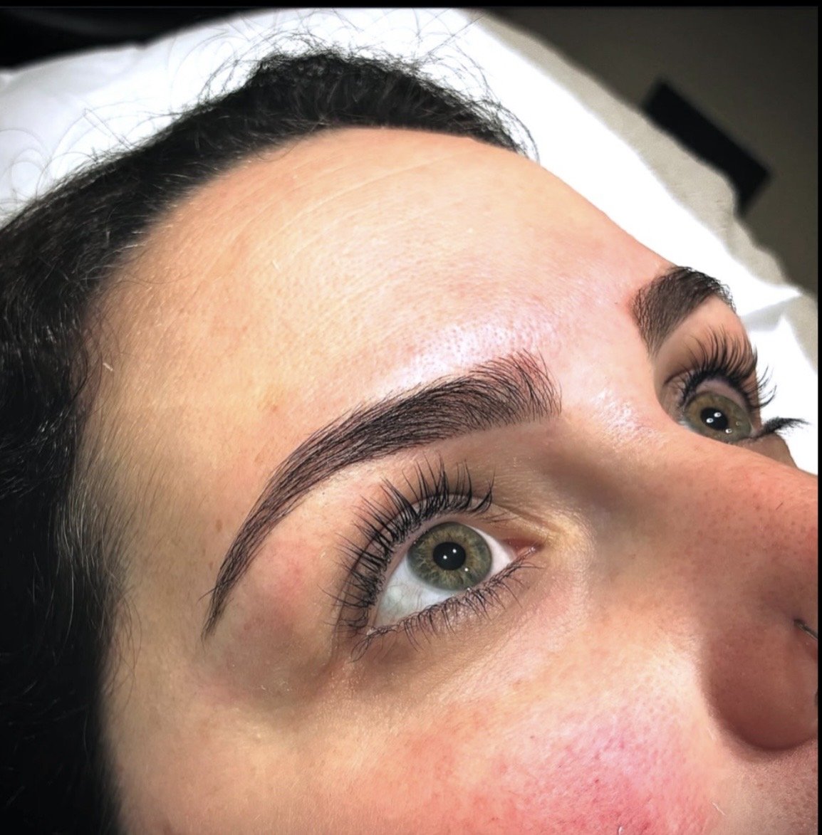 Close-up of a woman's face showing freshly done eyebrow microblading, with well-shaped, dark, and defined eyebrows, and long eyelashes.