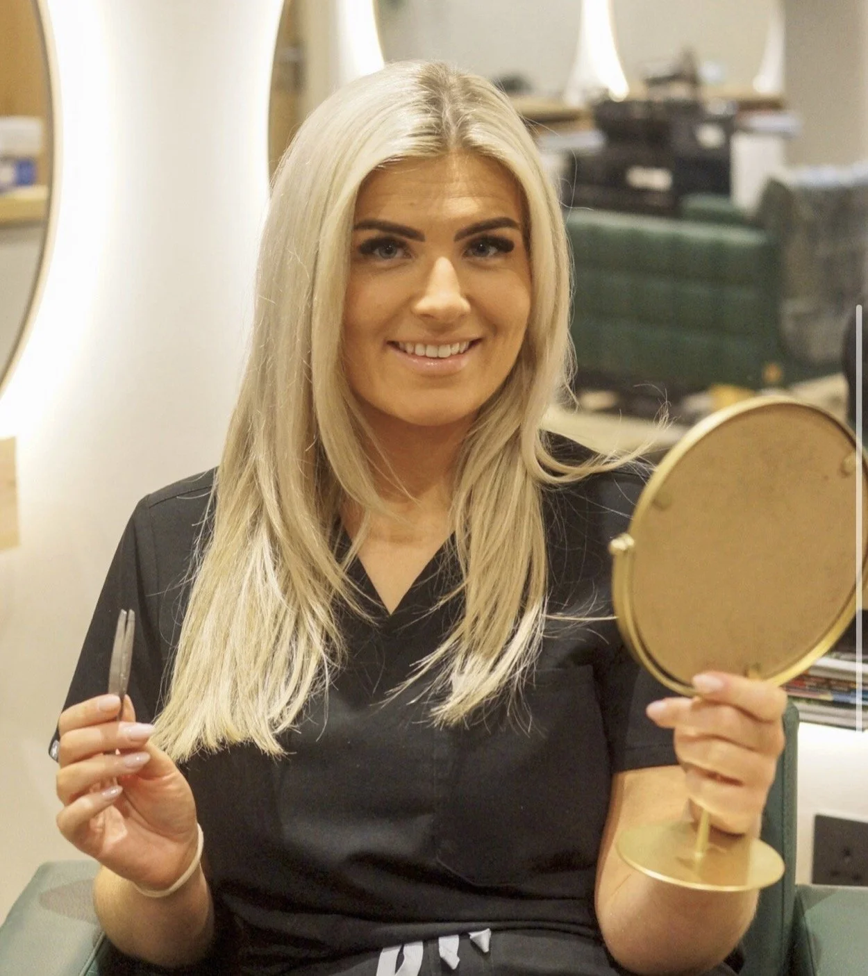 A woman with long blonde hair is sitting in front of a mirror, holding a small round mirror in her right hand and a pair of scissors in her left hand, smiling in a room with a background of shelves and stacked items.