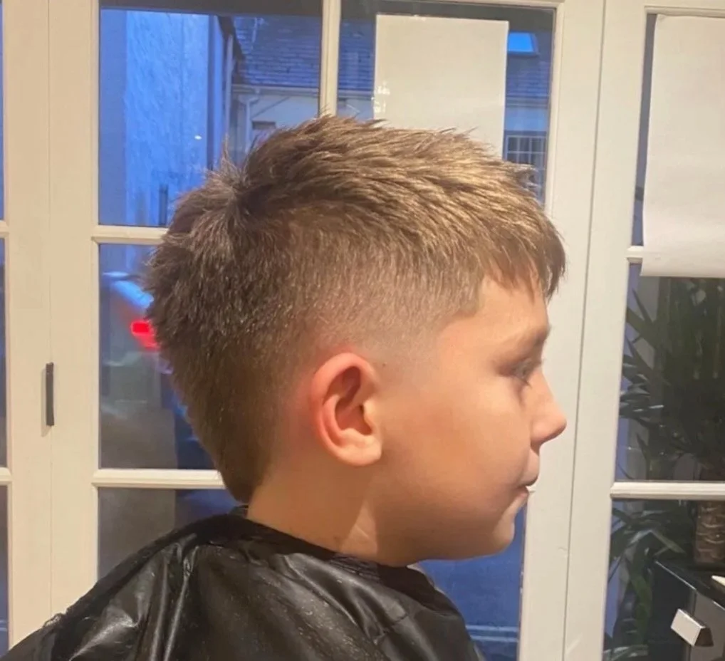 Side view of a young boy with a short haircut, sitting indoors in front of glass doors.