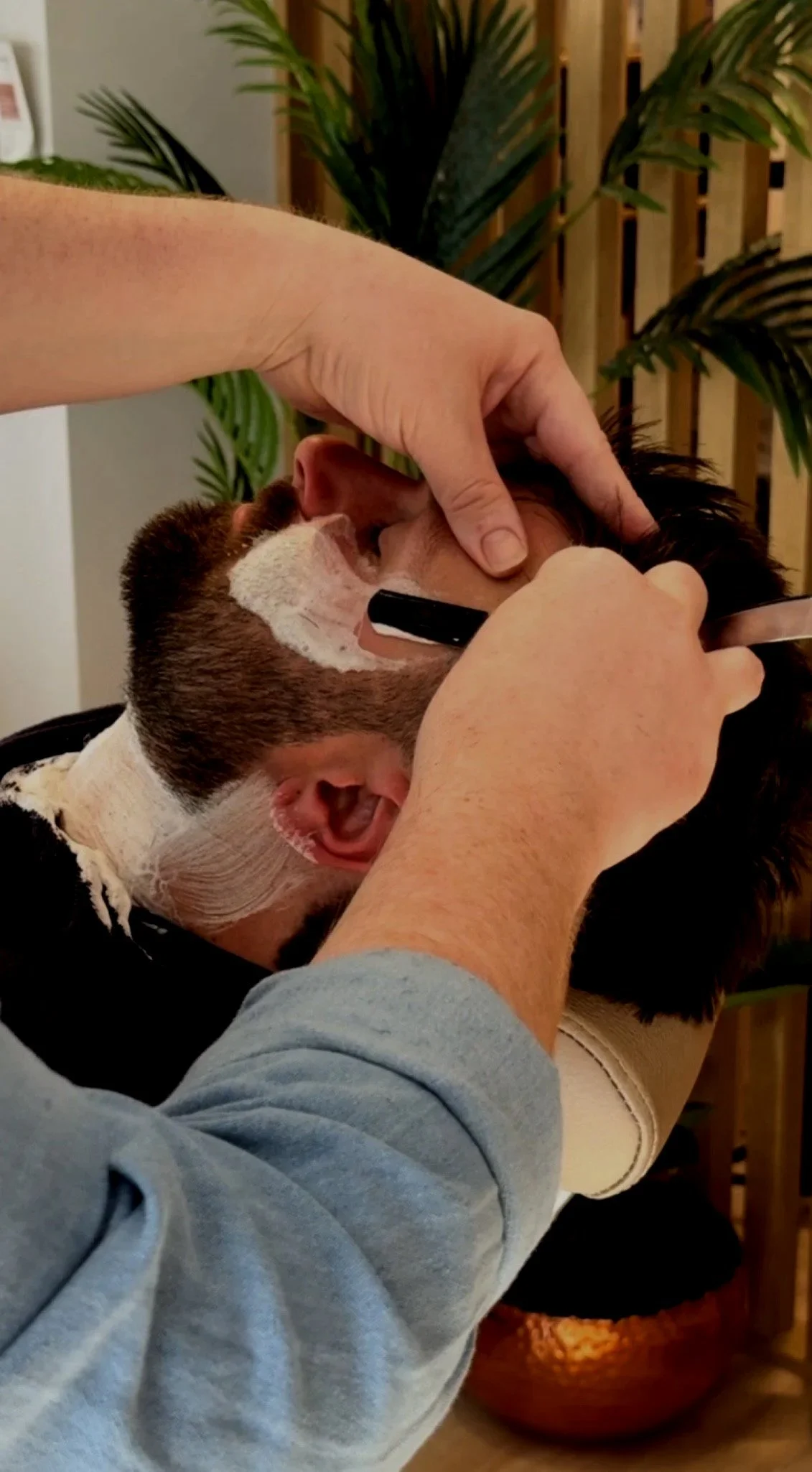 A person getting a haircut with soap and a razor near the beard while lying on a chair, with hands working on their face, and surrounded by plants and a wooden background.