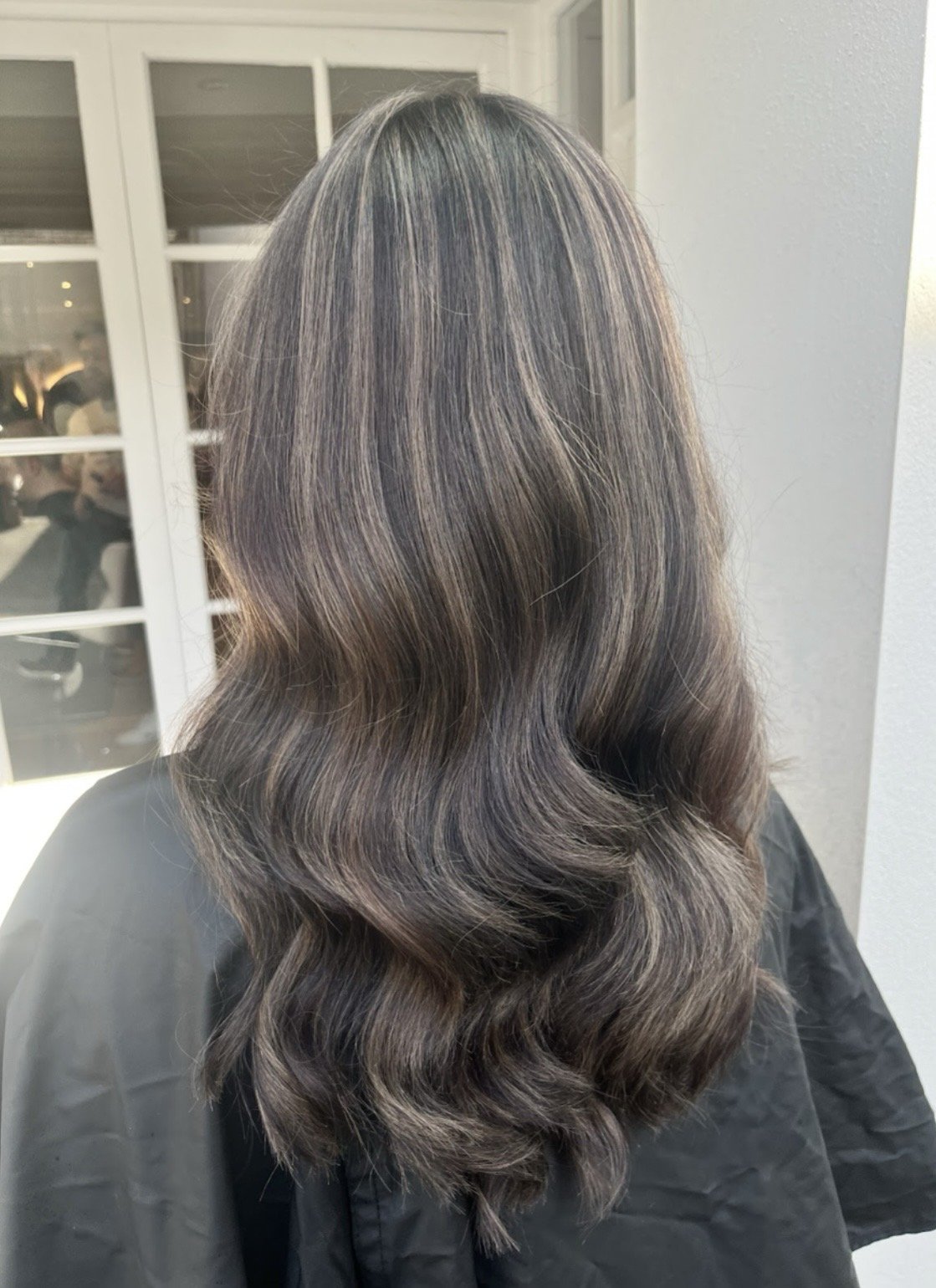 Back view of a woman with wavy, shoulder-length hair with highlights, standing indoors near a window.