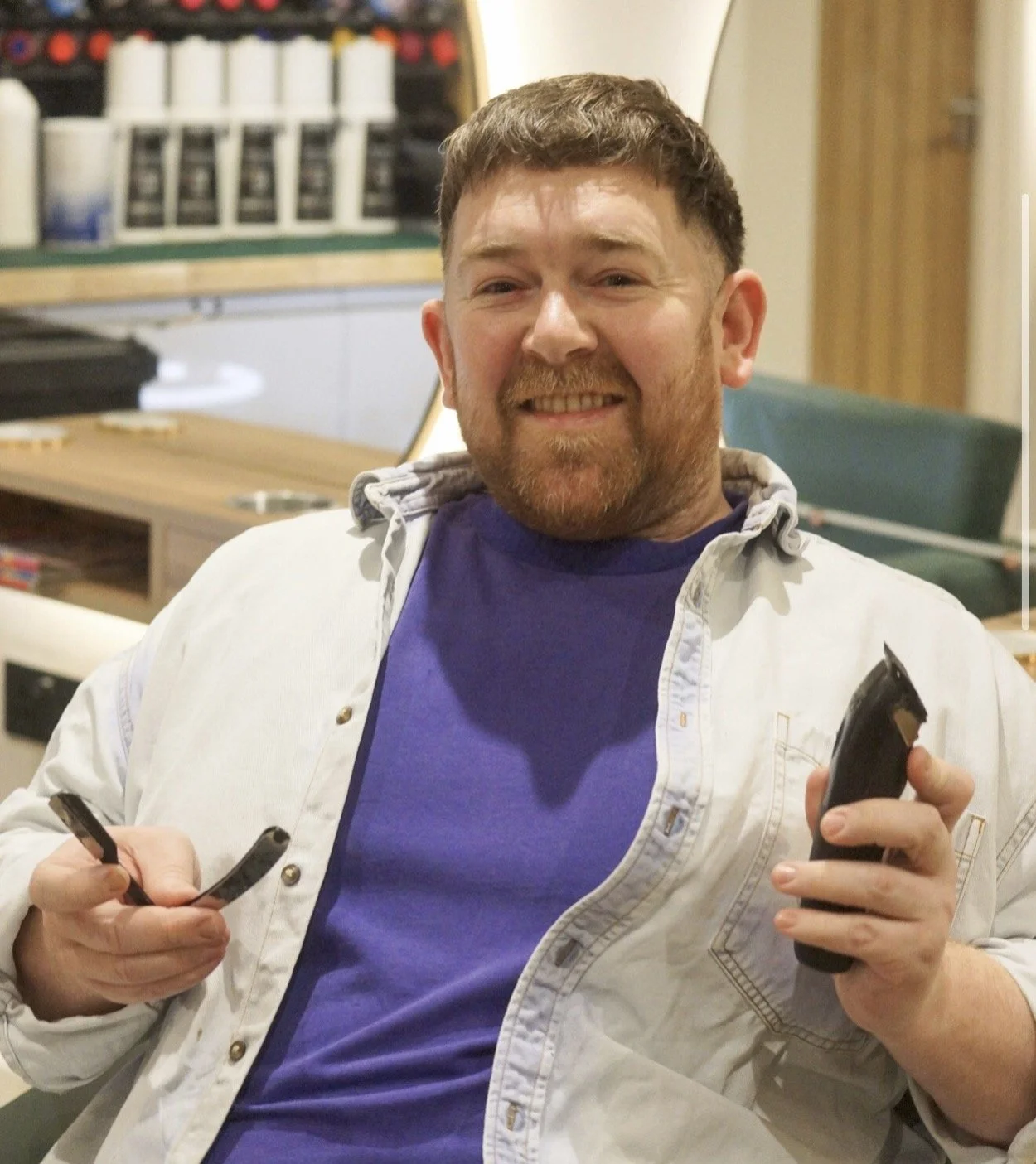 A man with short brown hair and a beard smiling, holding a phone in his right hand and a remote control in his left, sitting in a casual indoor setting.