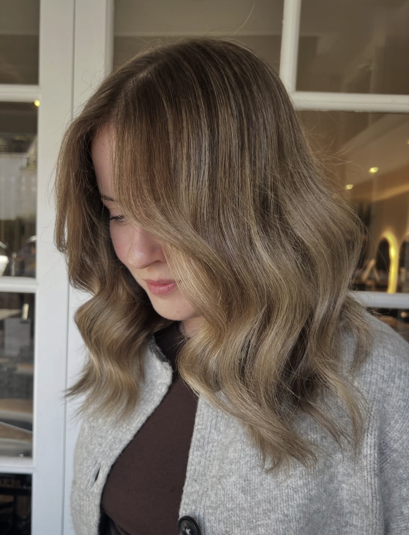 Woman with shoulder-length wavy light brown hair, looking down, wearing a gray cardigan and a dark top, standing indoors near glass doors.