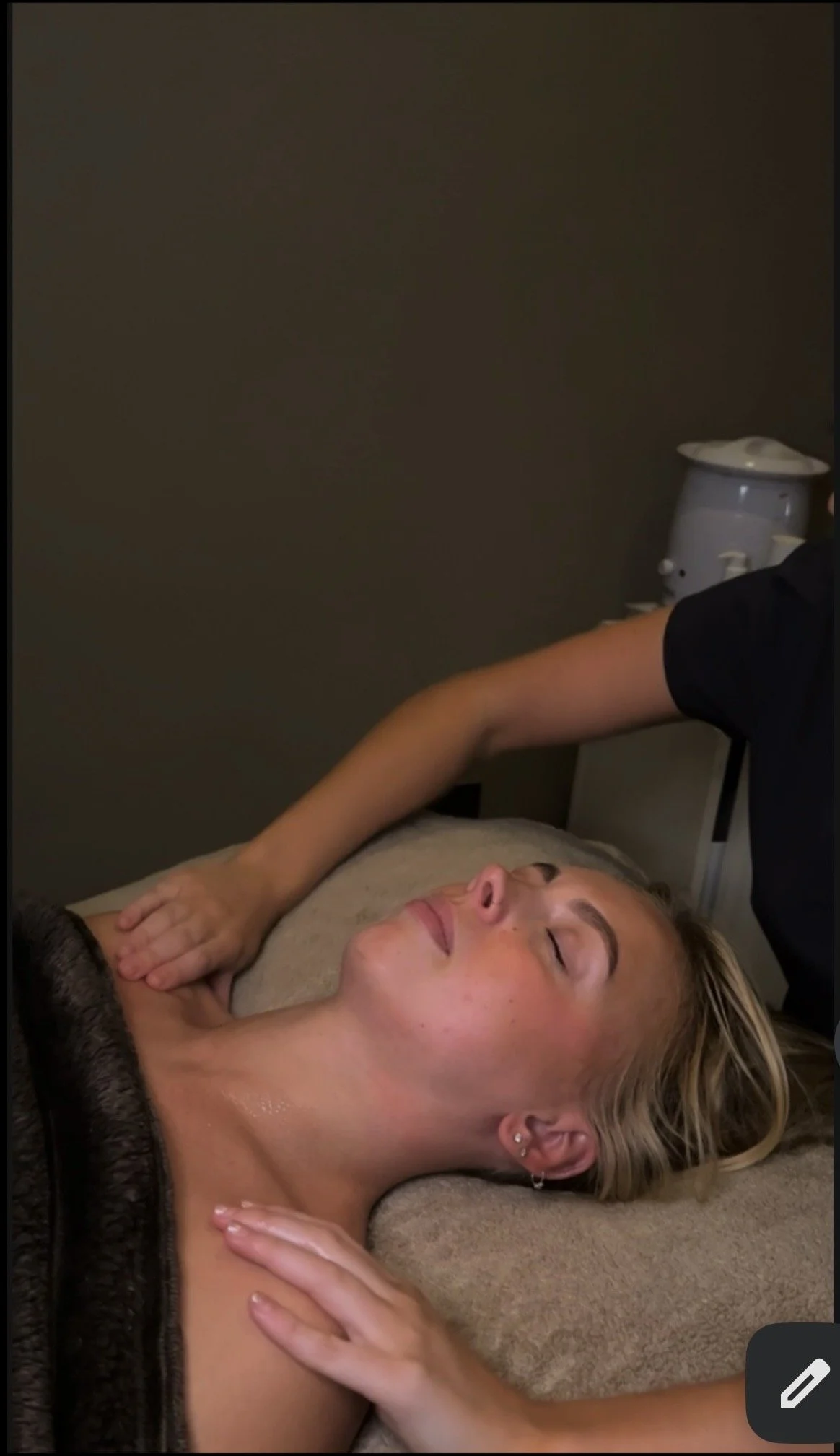 A woman lying on her back receiving a massage, with eyes closed and a peaceful expression, in a dimly lit room.