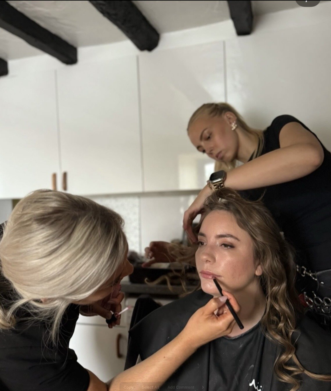 A woman with curly hair is sitting in a makeup chair receiving makeup application from a professional makeup artist, while another woman does her hair.