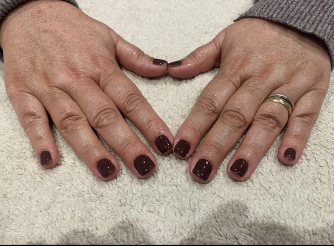 Two hands with dark red painted nails placed on a light-colored towel, forming a heart shape with the fingertips. One hand has a gold wedding band.