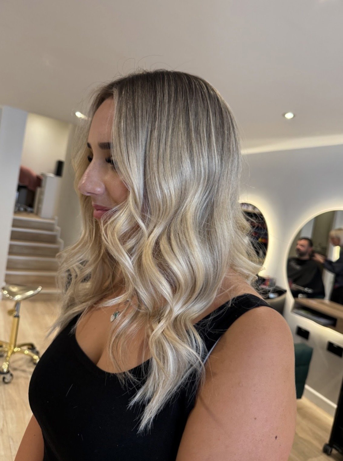 Side profile of a woman with long, wavy blonde hair, sitting in a salon with a stylist working in the background.