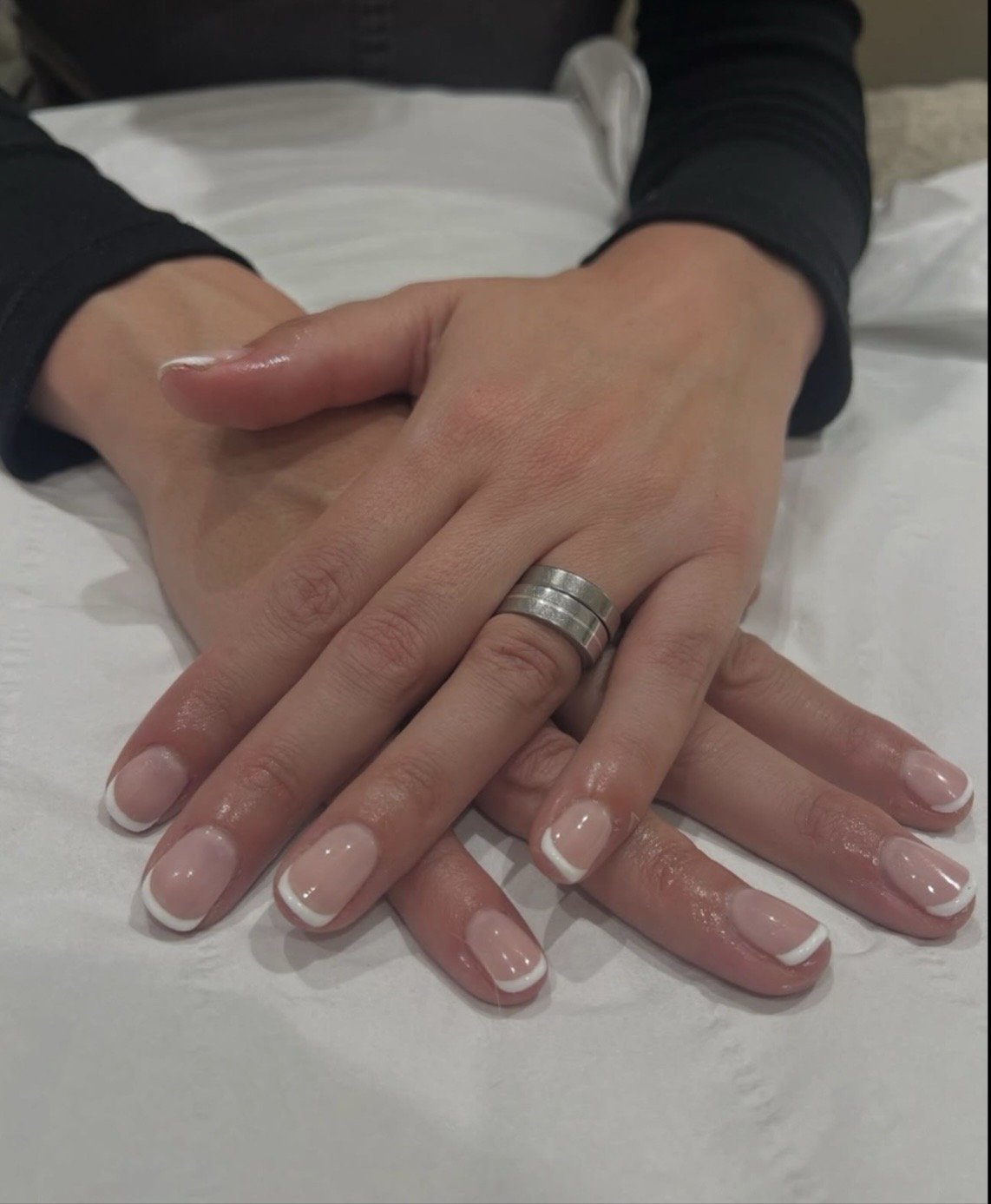 Close-up of two hands with manicured nails, one with a silver ring, resting on a white surface. The person is wearing a black long sleeve shirt.