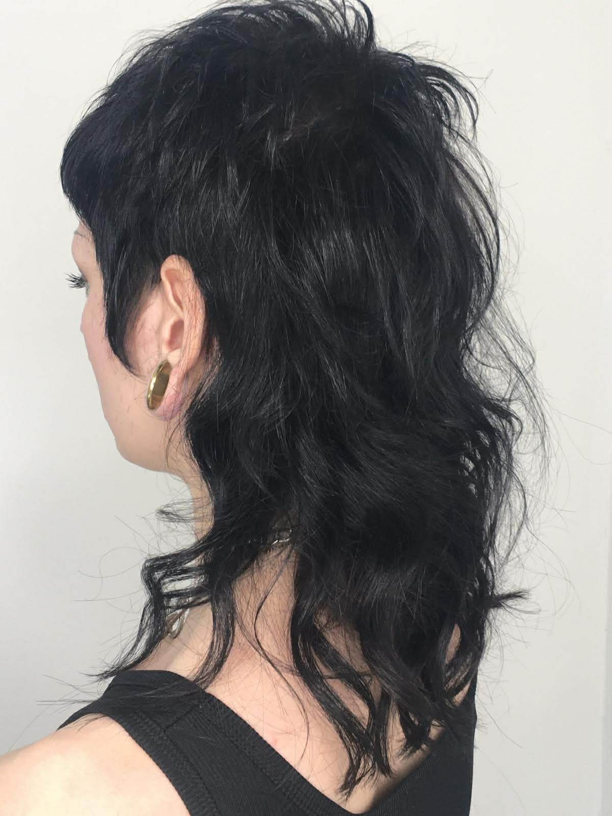 Side view of a woman with dark, wavy, layered hair wearing a black top and a gold earring.