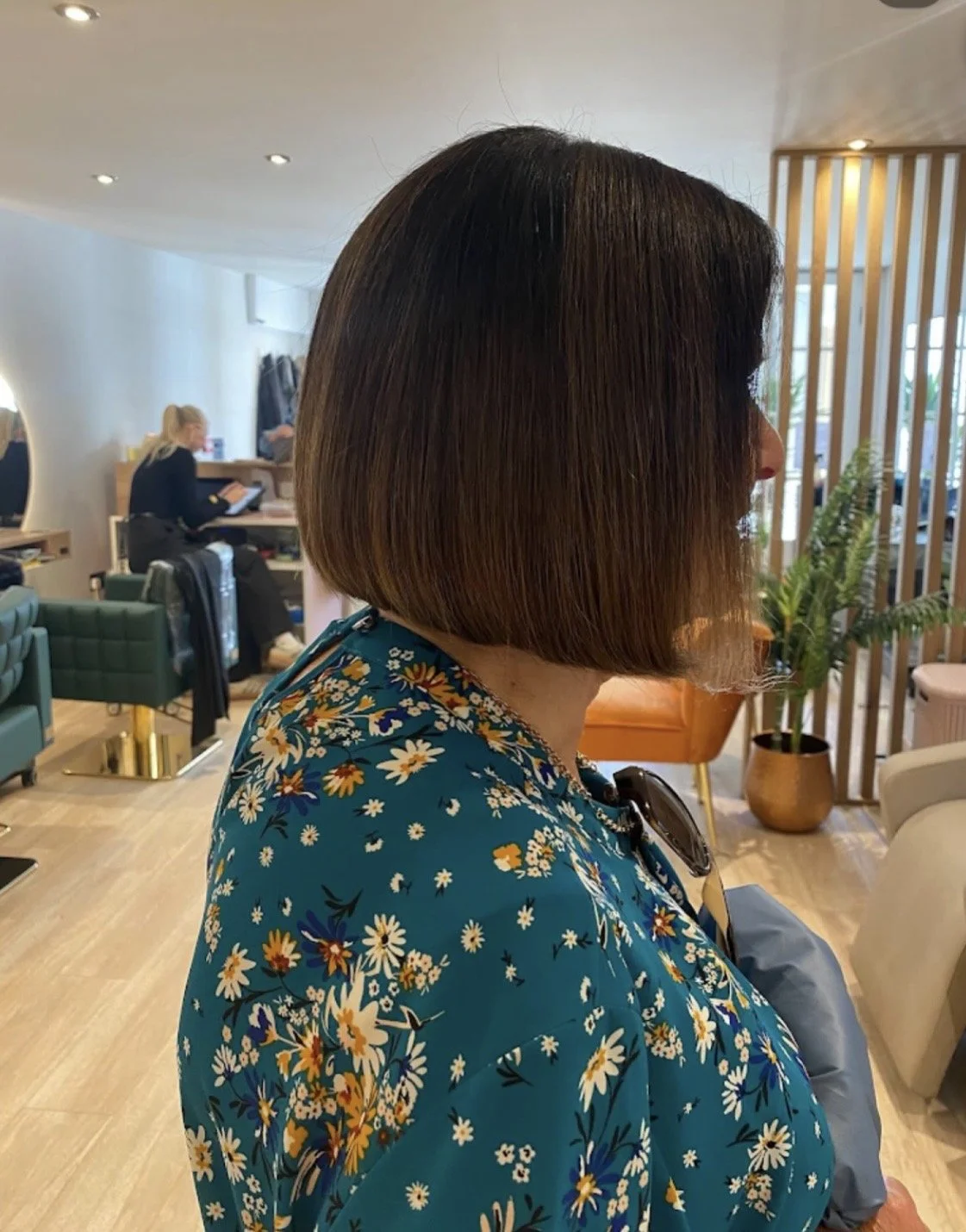 Side view of a woman with a short brown bob haircut, wearing a blue floral top inside a stylish salon or lounge with people and furniture in the background.