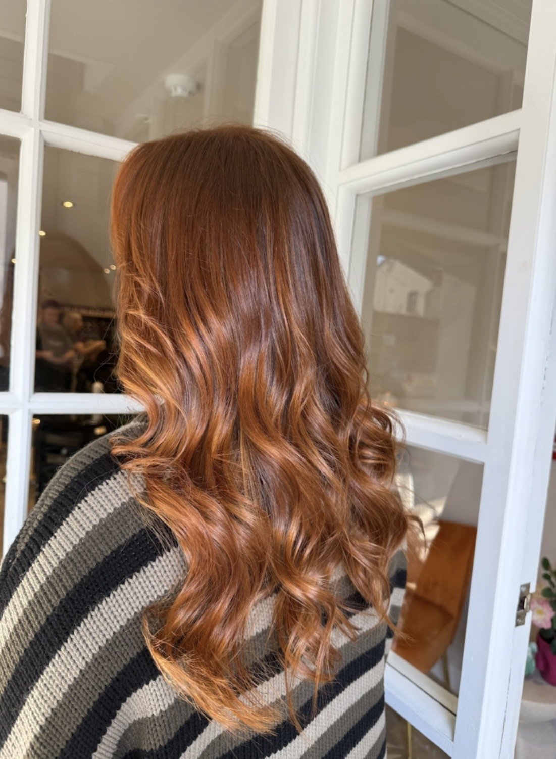 Back view of a woman with long, wavy, red hair, wearing a black and beige striped sweater.