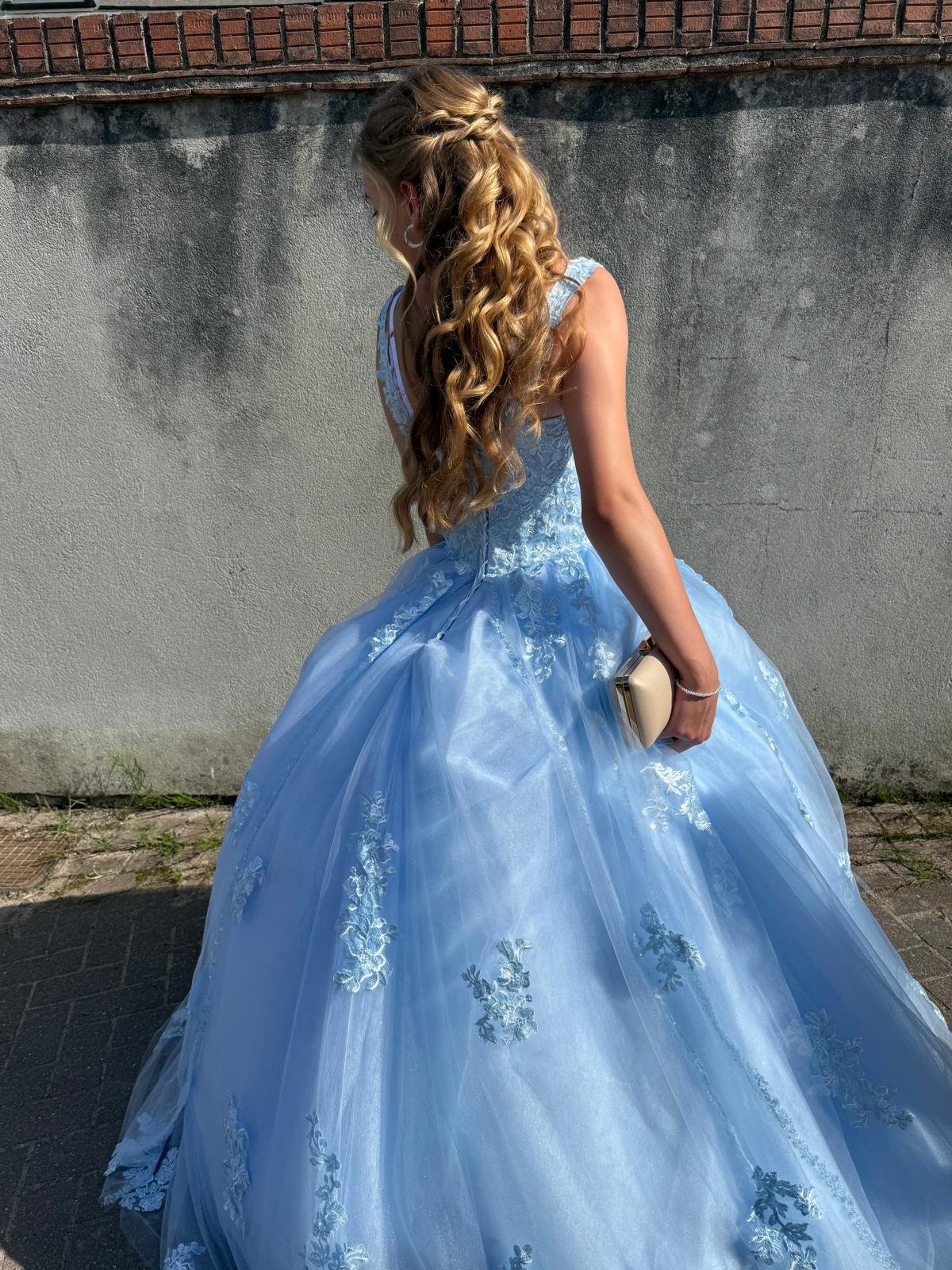 A young woman with long, wavy blonde hair wearing a light blue ball gown with floral embroidery, holding a beige clutch purse, standing outdoors against a concrete wall and brick surface.