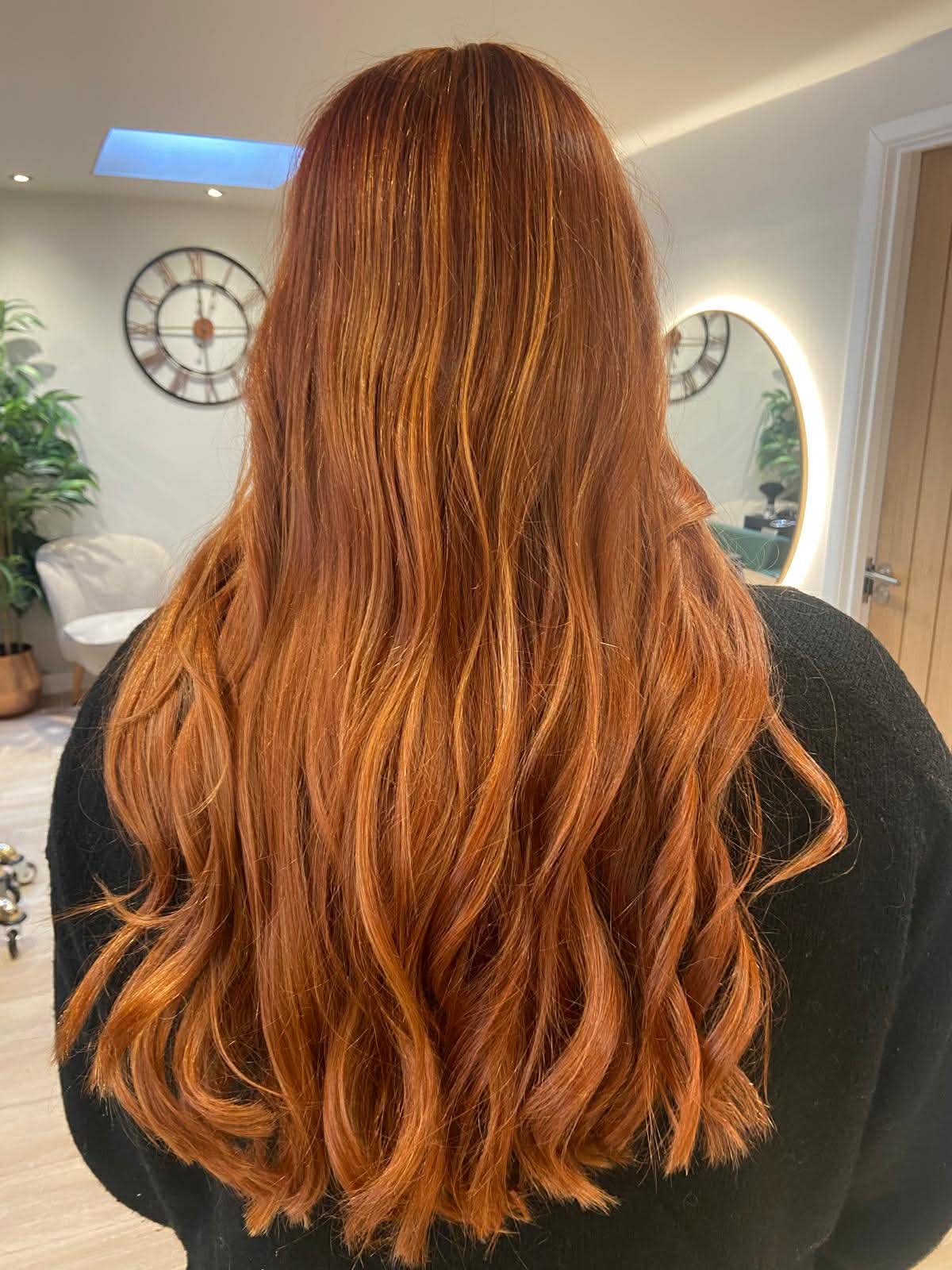 Back view of a woman with long, wavy, red hair in an indoor setting with a mirror and clock in the background.