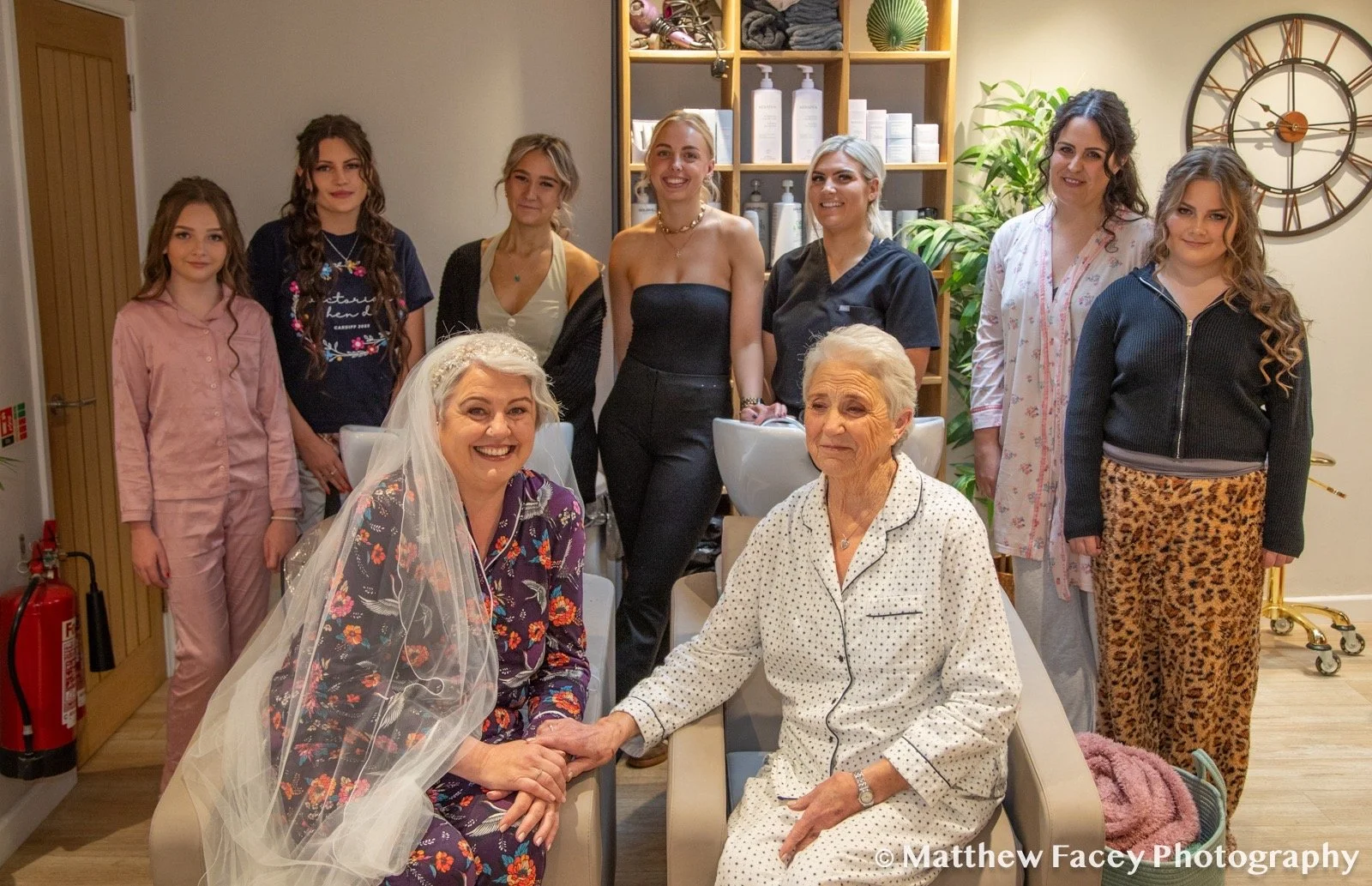 Group of women, including a bride with a veil and an older woman, posing in a salon with hair washing stations and a large clock on the wall.