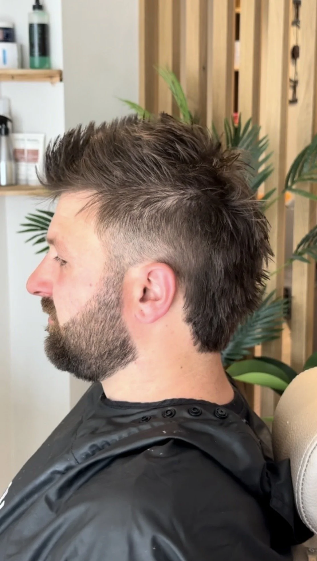 Side profile of a man with a fresh haircut and beard, sitting in a salon chair, with wooden wall and plants in the background.
