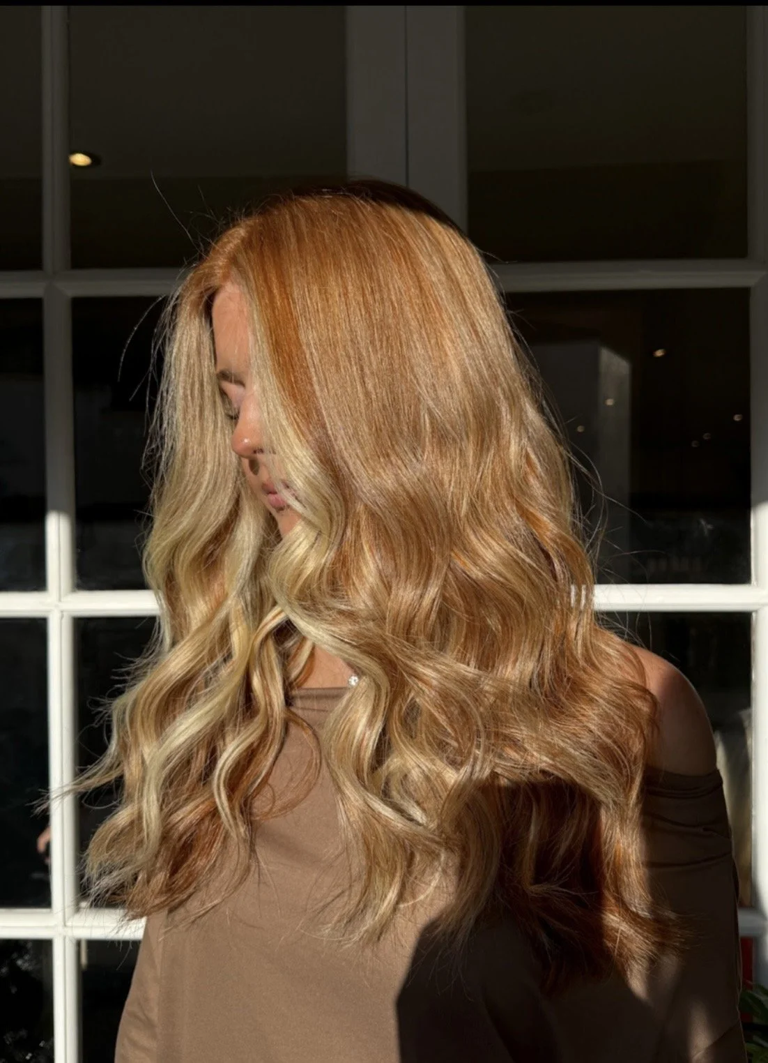 A woman with long, wavy, blonde hair standing in front of a window, looking down, with soft light illuminating her hair.