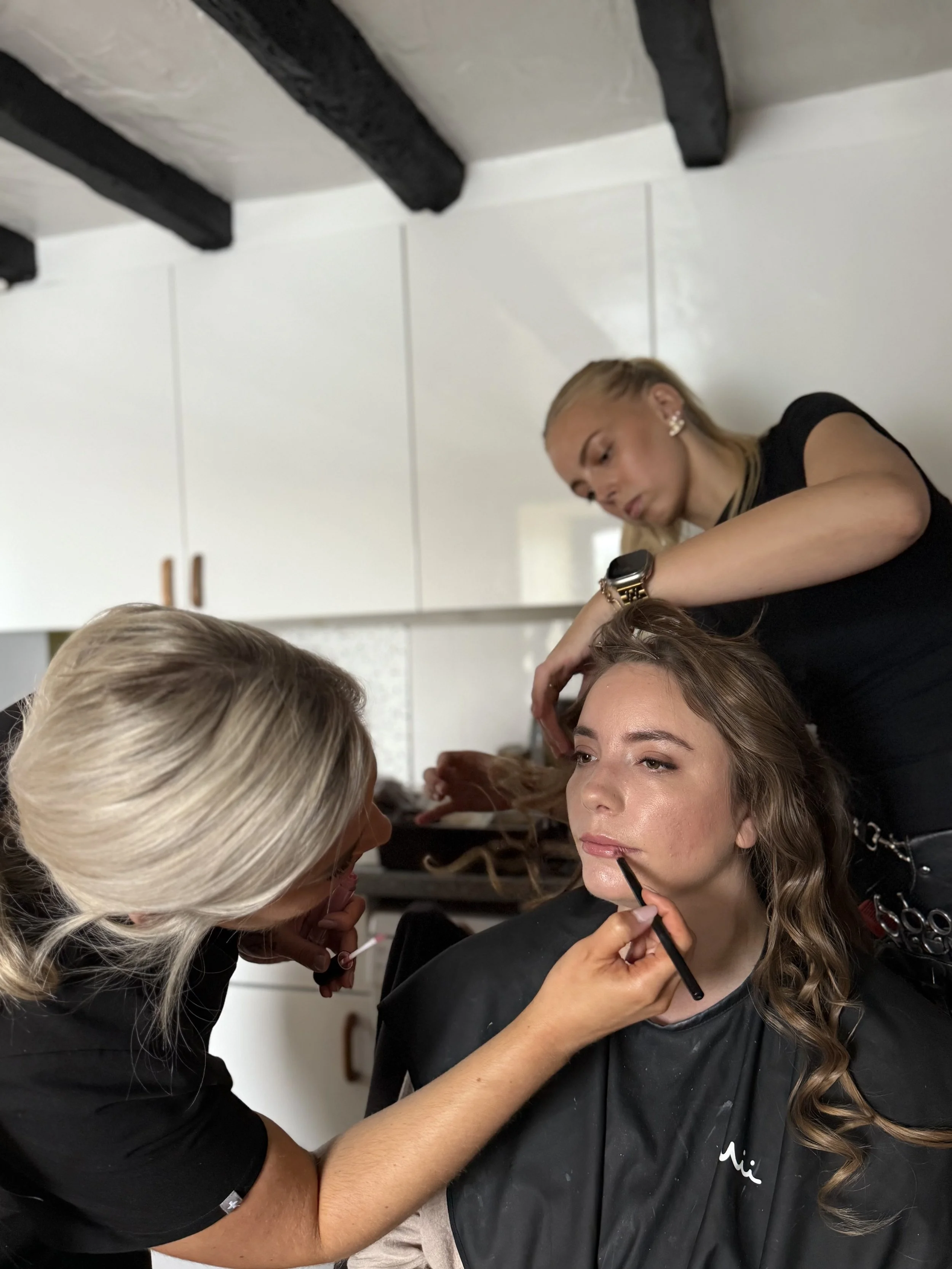 A woman with long, wavy brown hair is having her makeup done by a makeup artist, while another stylist works on her hair in a salon or backstage setting.
