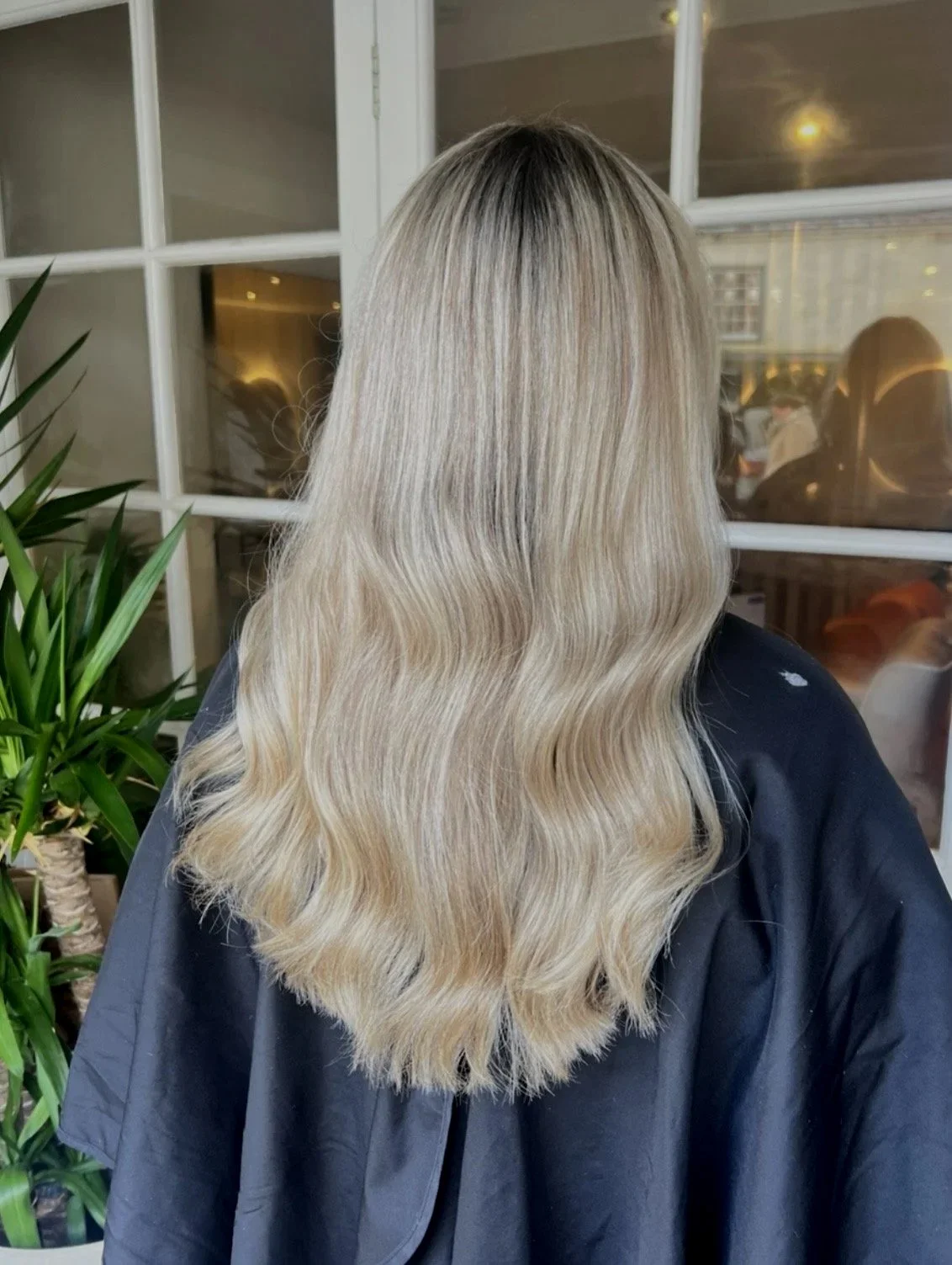 Back of a woman with long, wavy blonde hair sitting in a salon, wearing a black cape, with a plant and windows in the background.