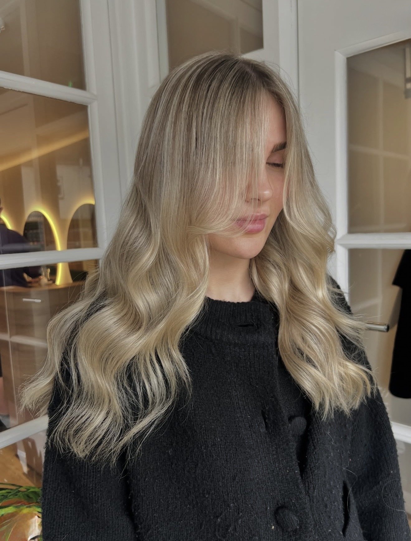A woman with blonde, wavy hair wearing a black sweater, standing indoors near glass-paneled doors.