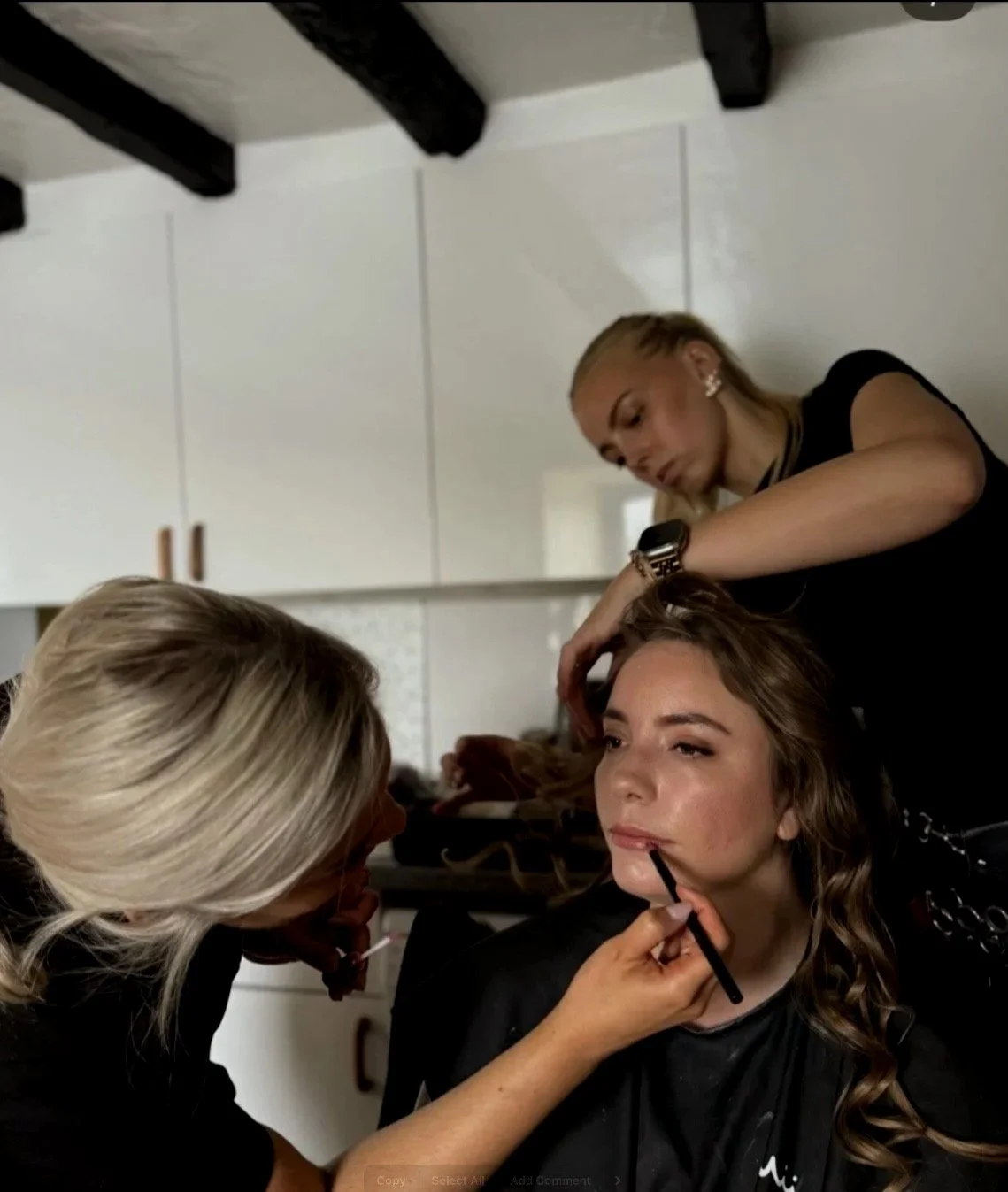Makeup artist applying makeup on a woman with long, curly hair while another woman with blonde hair watches and styles her hair.