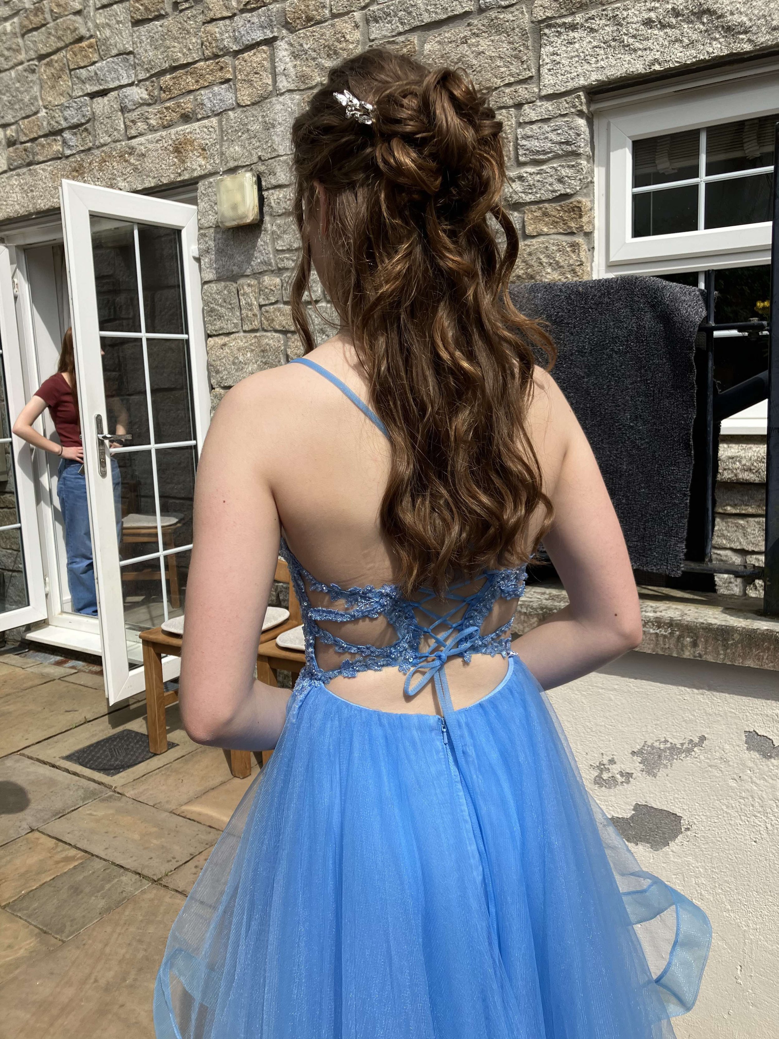 Young woman in a blue dress with a lace-up back, long wavy hair, standing outside near a stone wall and open window. She is facing away from the camera.