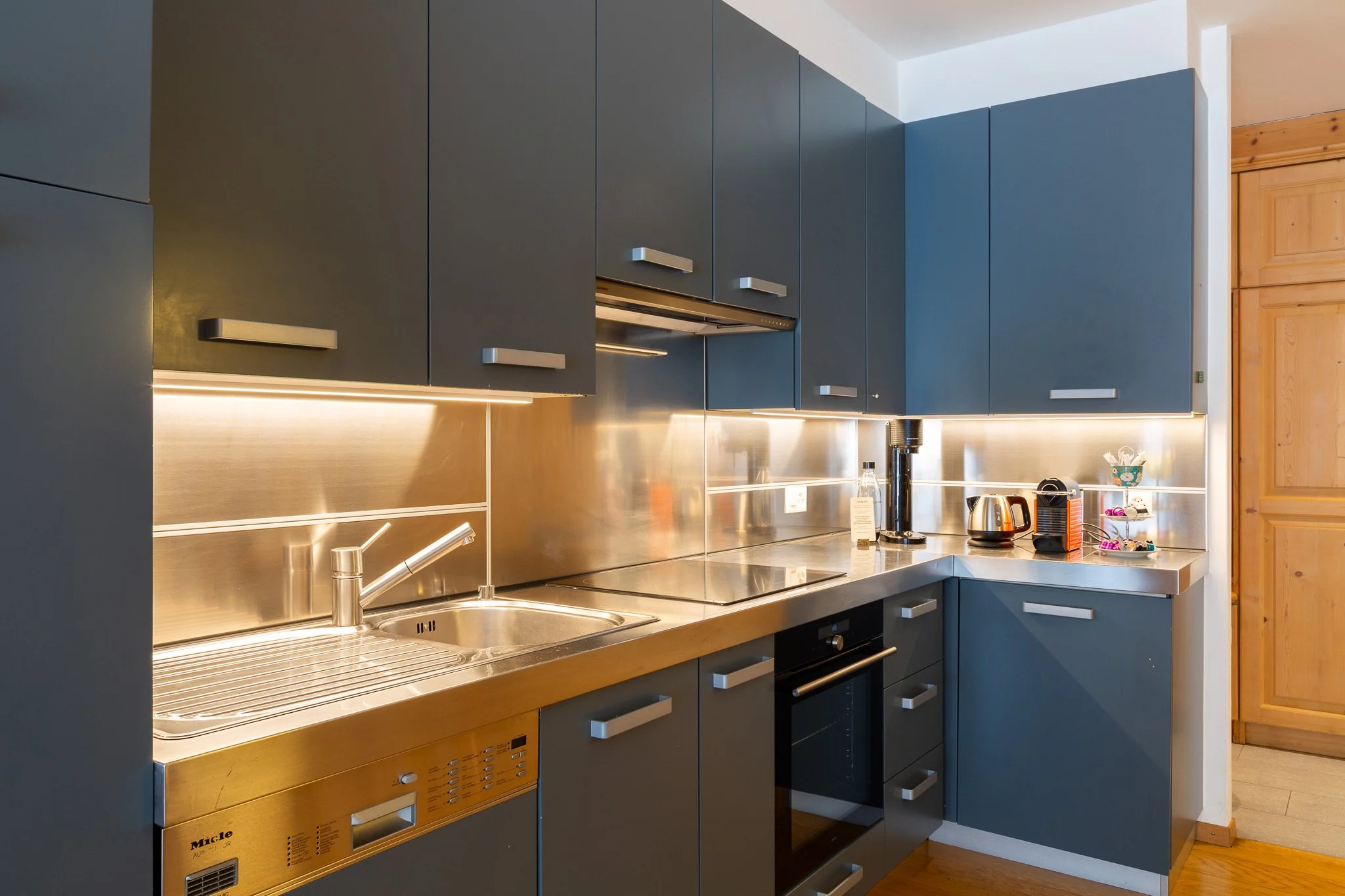 Modern kitchen with blue cabinets, stainless steel countertop and backsplash, small appliances, and wooden door.